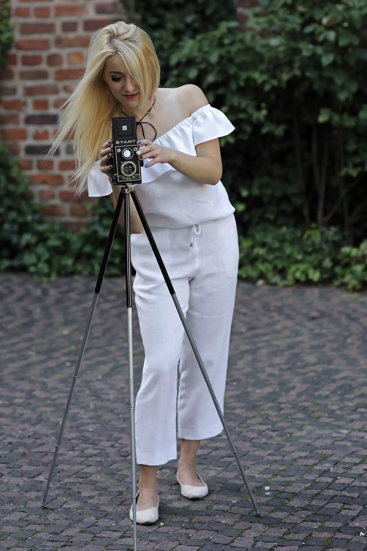 Blondynka stoi w lekkim rozkroku, pochylona nad starym aparatem fotograficznym na trójnogu. Ma na sobie biały, lniany komplet: bluzkę hiszpankę i spodnie Mira o długości 7/8. Stylizacja jest letnia i artystyczna.