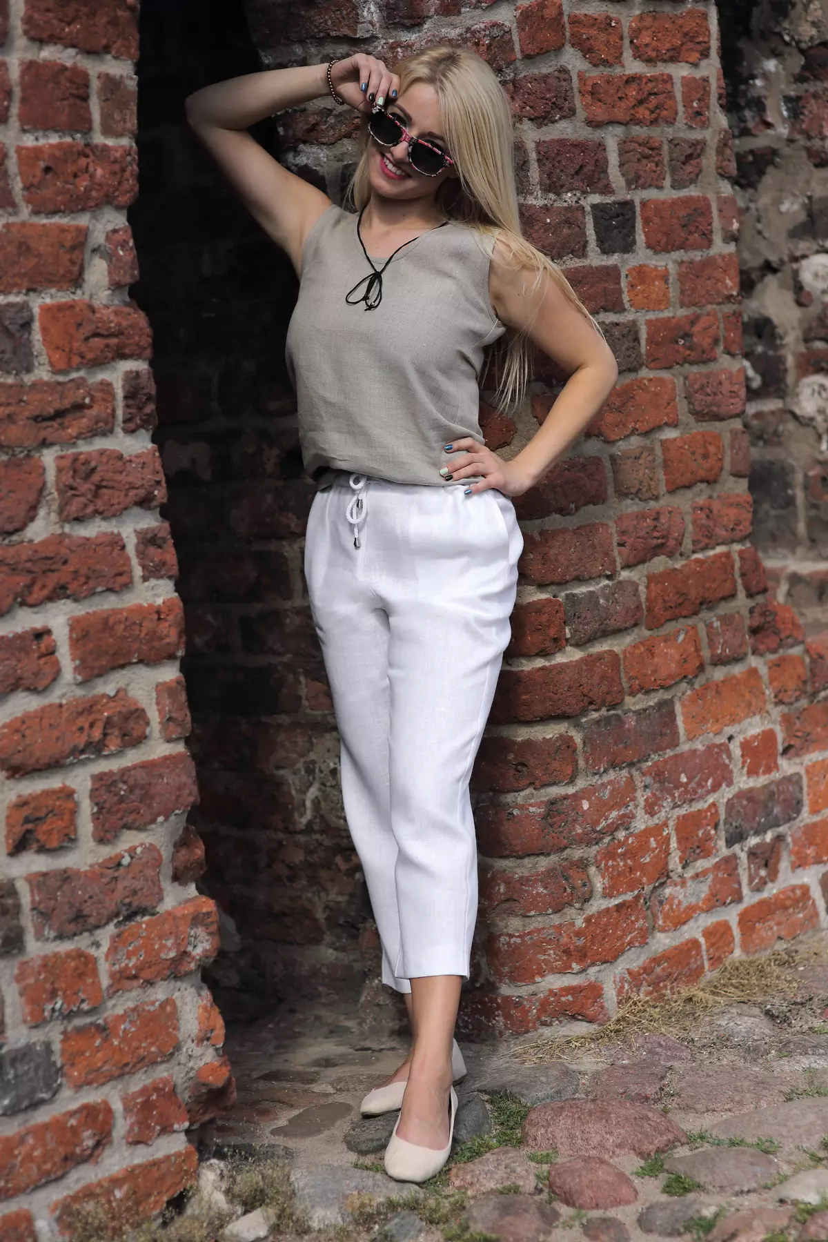 Uśmiechnięta blondynka w okularach przeciwsłonecznych opiera się o ceglany mur. Ma na sobie białe spodnie Mira i beżowy top. Spodnie są luźne w biodrach i lekko zwężają się ku dołowi, kończąc się nad kostką.