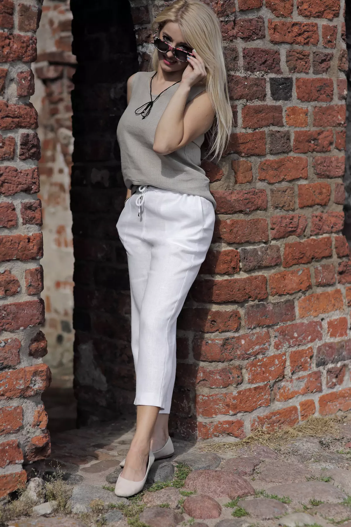 Modelka stoi w ceglanym łuku, poprawiając okulary. Białe spodnie Mira kontrastują z surową cegłą. Luźny krój i długość 7/8 sprawiają, że spodnie wyglądają na bardzo przewiewne i komfortowe.