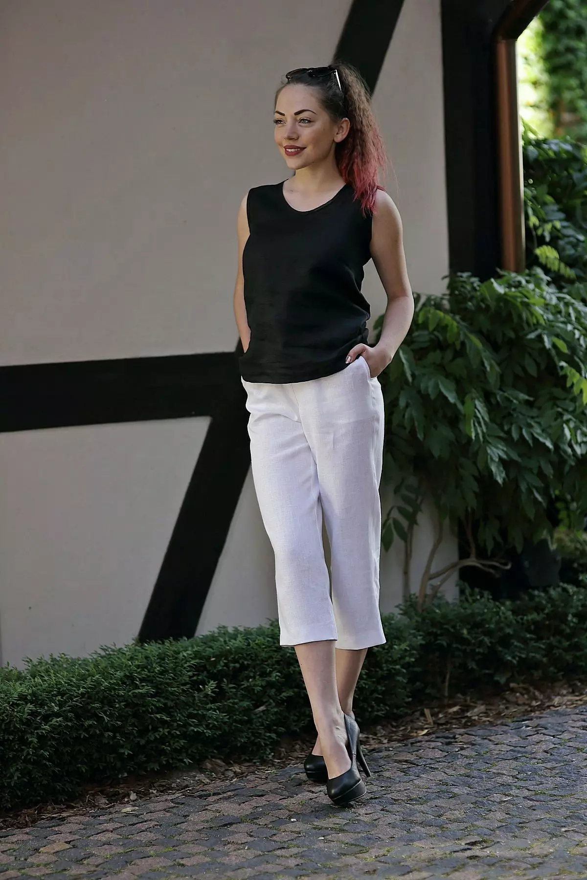 Brunetka stoi na brukowanej drodze. Ma na sobie białe spodnie Mira, czarny top bez rękawów i czarne buty na wysokim obcasie. Stylizacja pokazuje, że lniane rybaczki można nosić również w bardziej eleganckim wydaniu.
