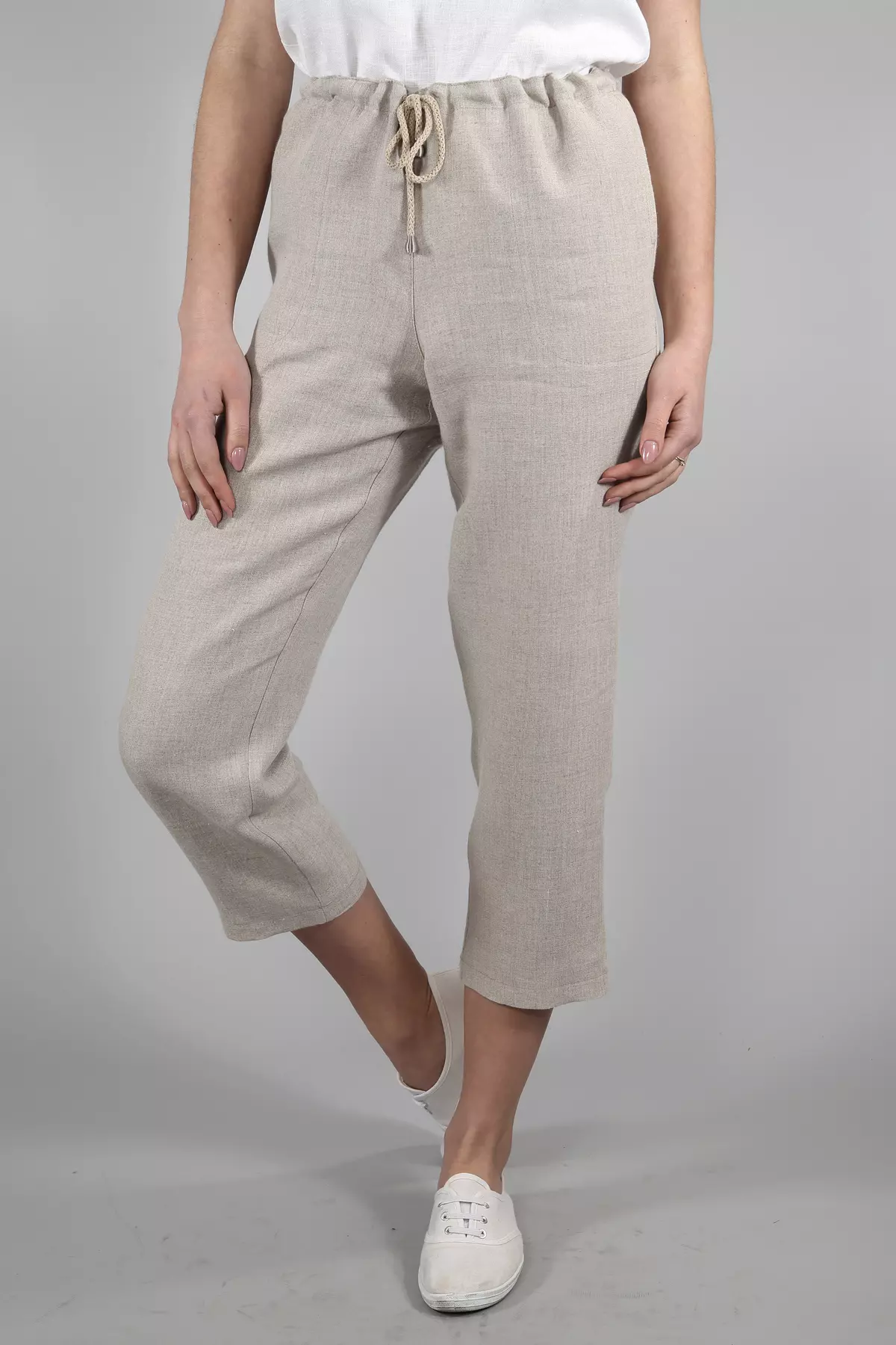 Modelka stoi na szarym tle, ma ugiętą prawą nogę. Ma na sobie spodnie Mira w kolorze naturalnym i biały top. Ręce trzyma luźno wzdłuż ciała. Spodnie wyglądają na bardzo wygodne i przewiewne.