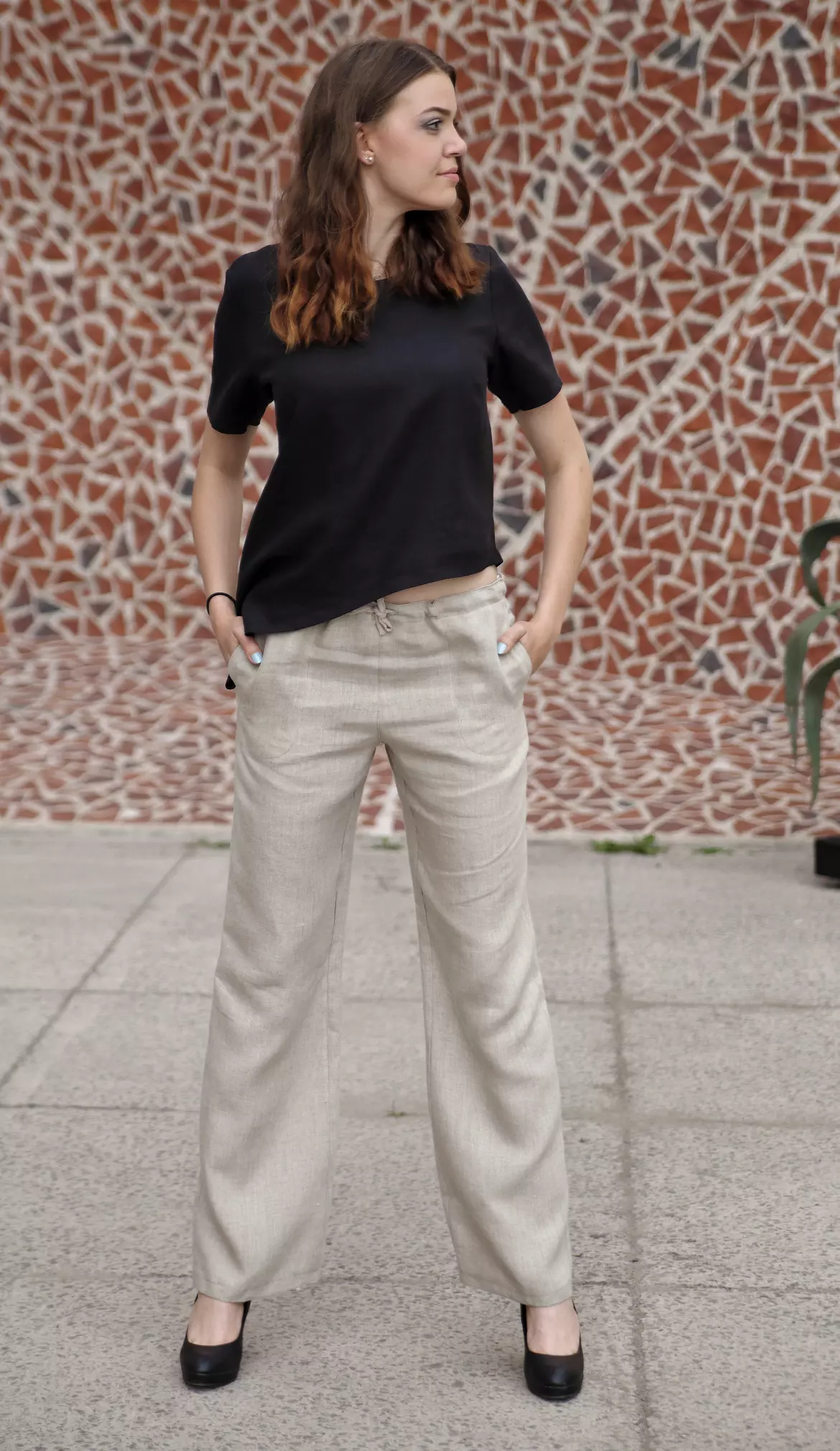 Modelka stoi na tle ściany z mozaiki. Dłonie trzyma w kieszeniach bocznych spodni. Spodnie Kaja mają widoczny sznurek w pasie i są wykonane z niebarwionego lnu. Stylizacja jest codzienna i wygodna.