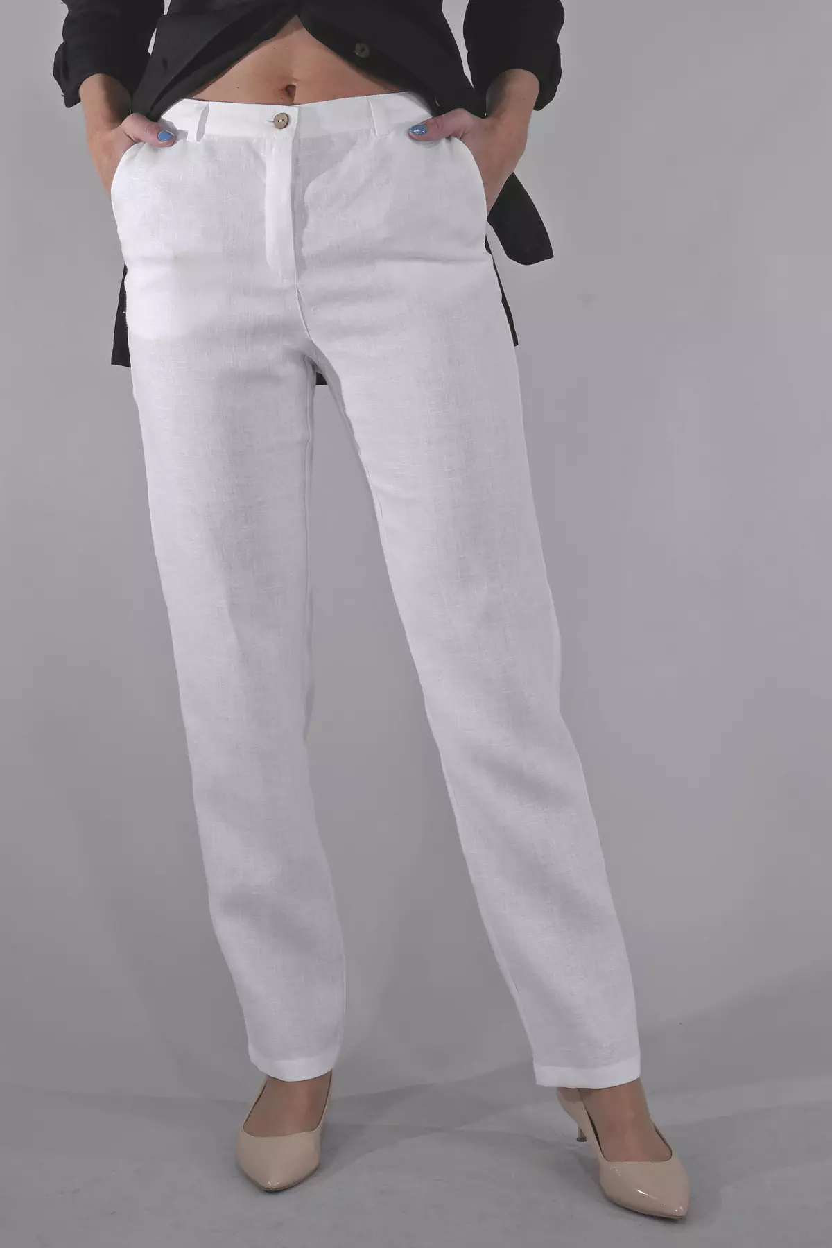 Modelka stoi przodem, prezentując białe spodnie lniane w pełnej okazałości. Spodnie mają klasyczny stan, zapięcie na guzik i rozporek oraz szlufki na pasek. Stylizacja zestawiona jest z kontrastową czarną koszulą i beżowymi butami.