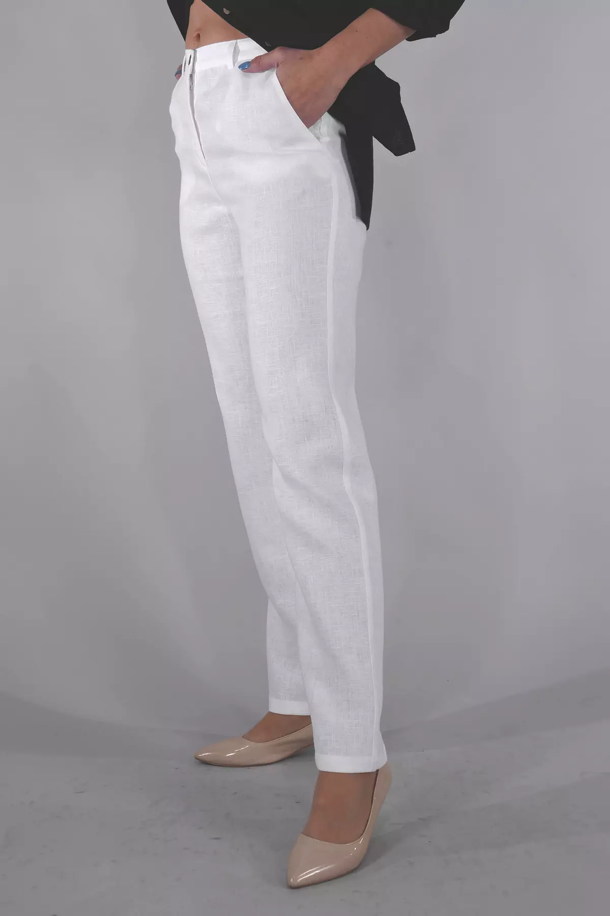 Kobieta stoi w półprofilu na szarym tle. Ma na sobie białe, lniane spodnie Kadi o prostym kroju. Lewą rękę trzyma w kieszeni bocznej. Spodnie są zapinane na guzik i mają szlufki w pasie. Modelka ma na sobie czarną koszulę.