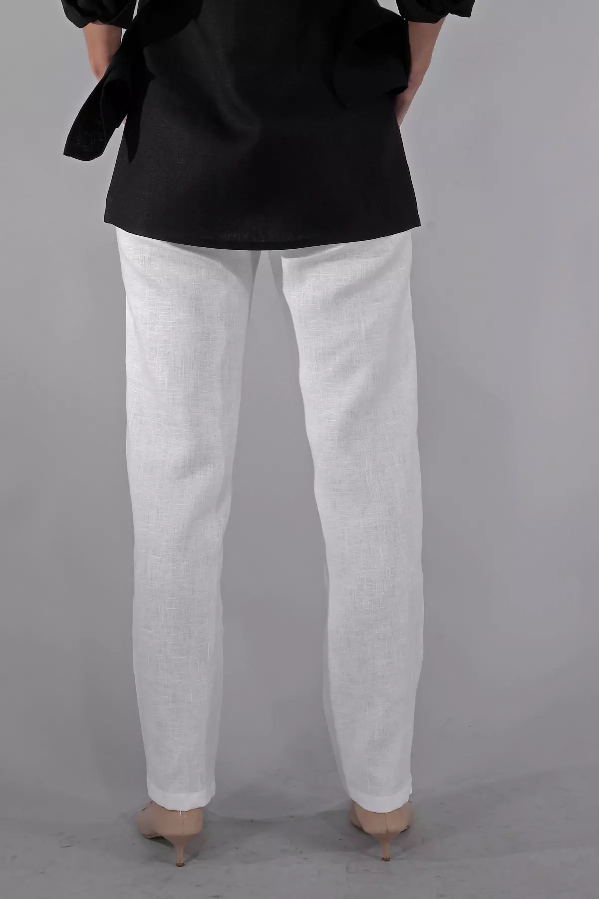 Widok spodni od tyłu. Nogawki są proste, lekko zwężane ku dołowi, nie opinają łydek. Biały len ma widoczną, naturalną strukturę. Modelka ma na sobie beżowe szpilki, które pasują do jasnych spodni.