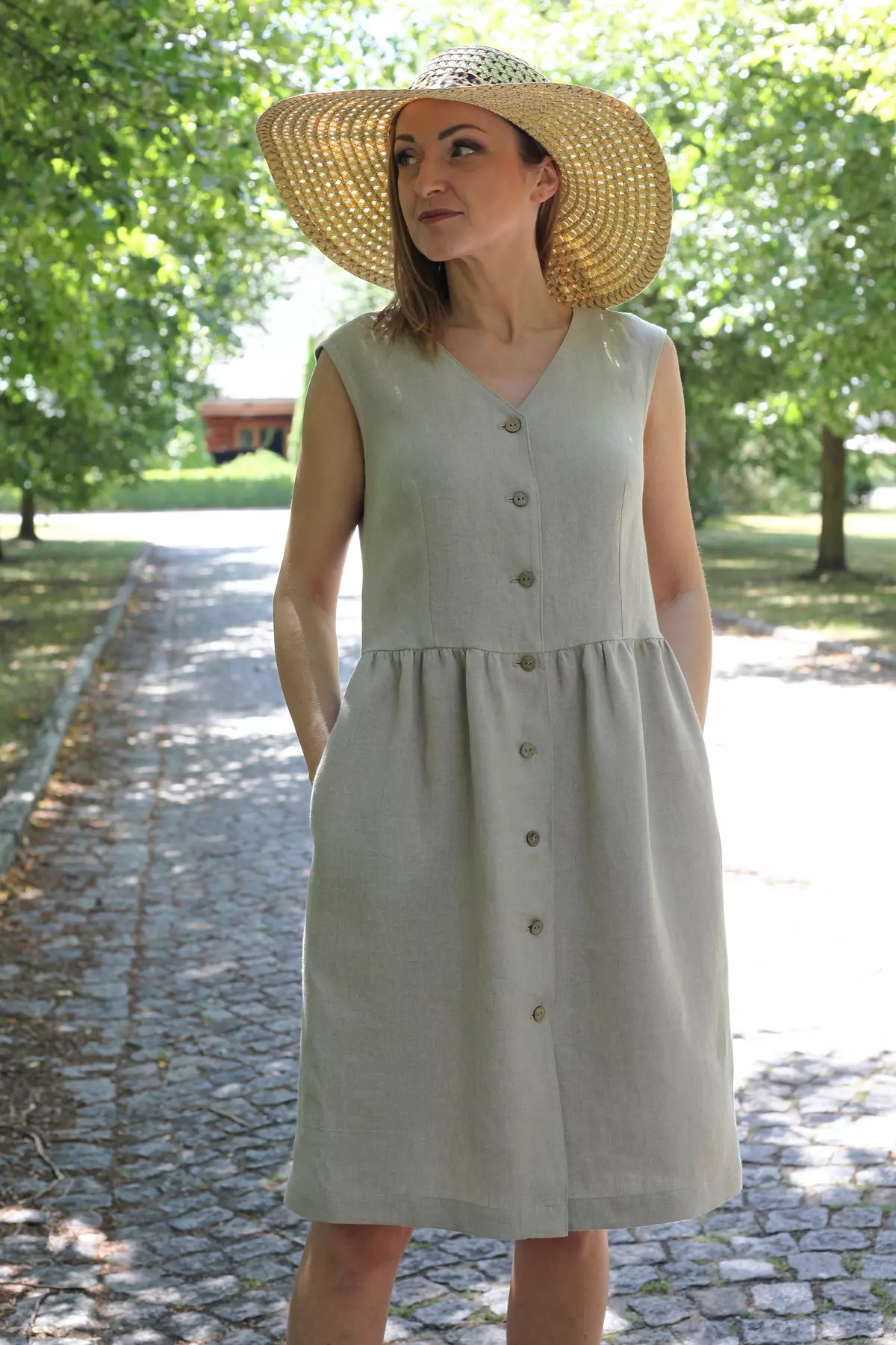 Modelka w beżowej, lnianej sukience Lucy bez rękawów, zapinanej na kokosowe guziki, stoi na ścieżce wśród drzew.