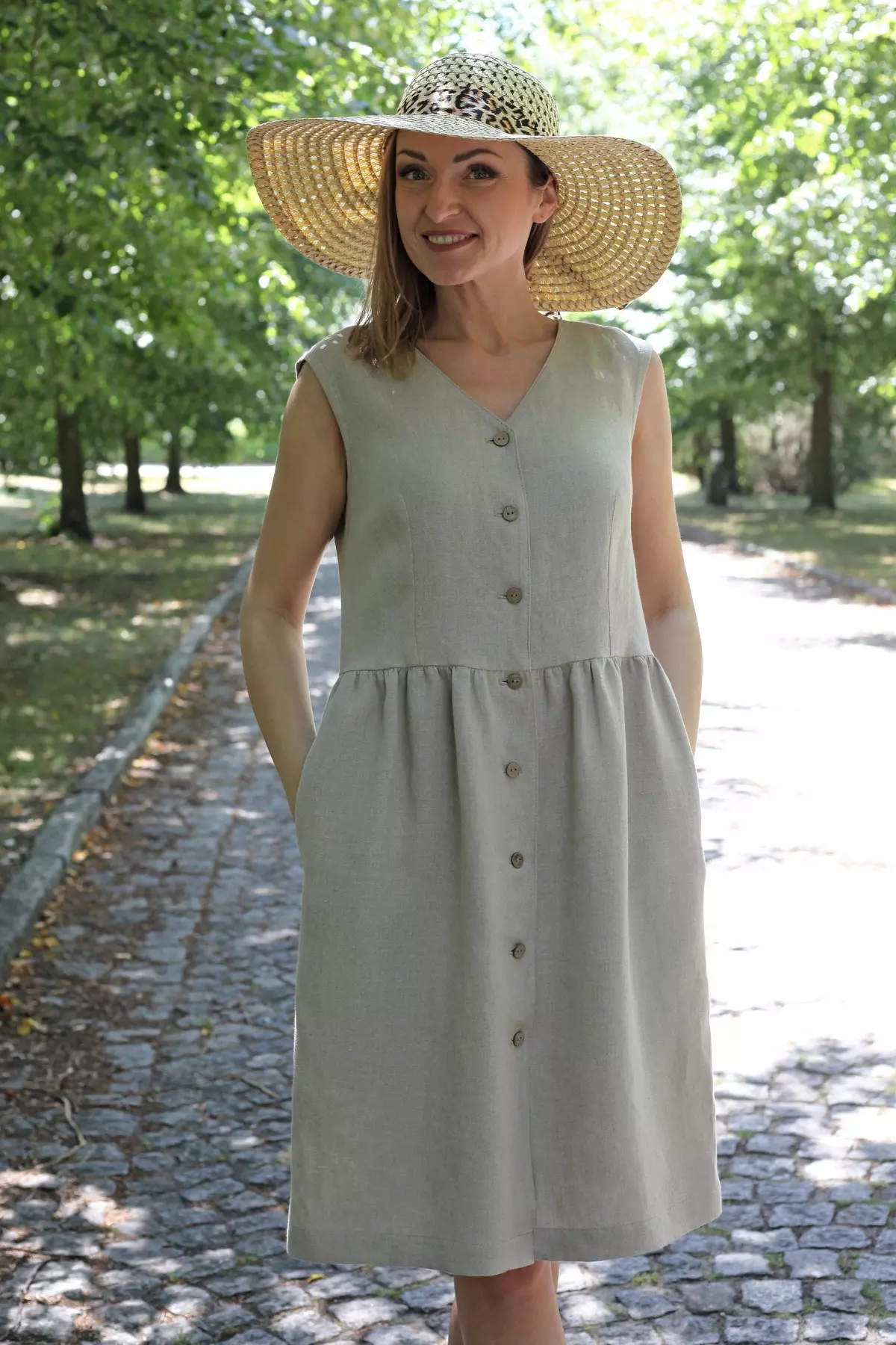 Modelka w lnianej sukience Lucy uśmiecha się, stojąc na słonecznej alejce w otoczeniu drzew, w kapeluszu ze słomy.