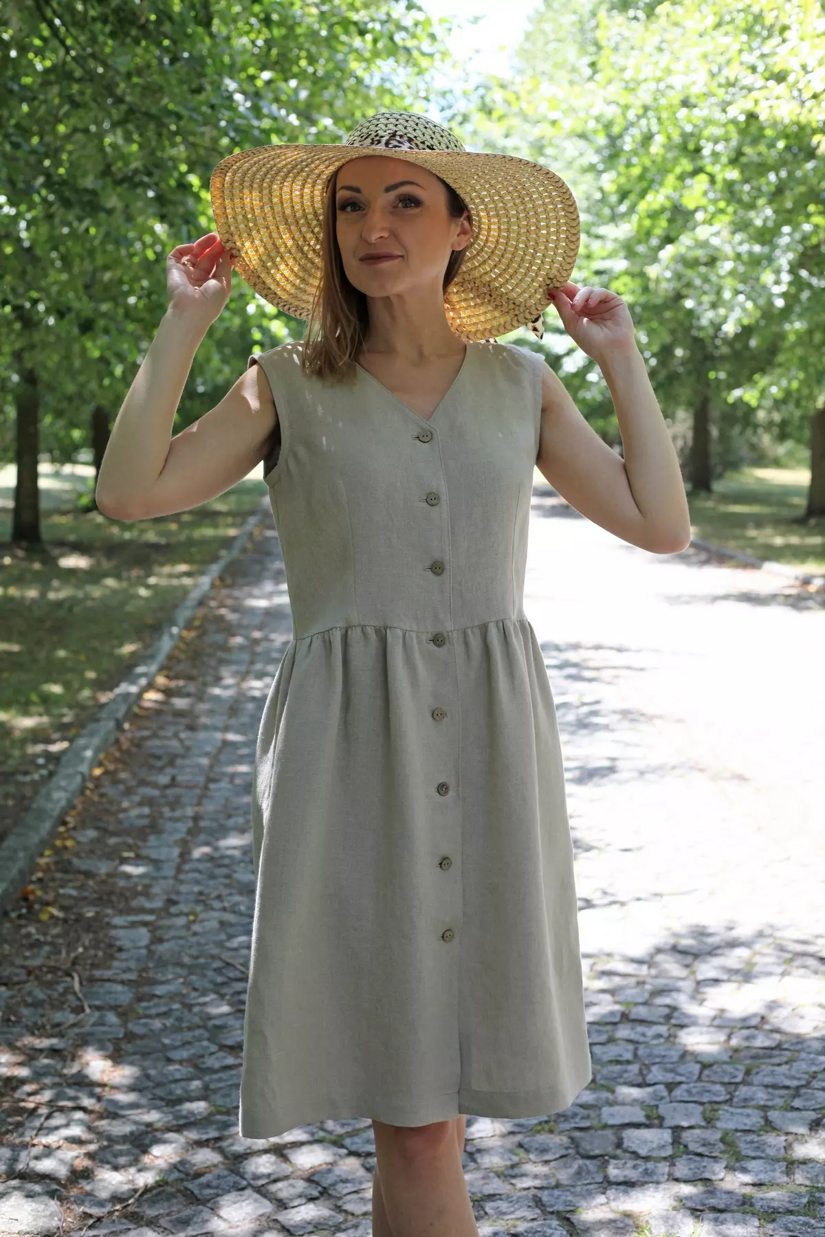 Modelka w słomkowym kapeluszu i lnianej sukience Lucy pozuje na kamiennej alejce w parku.