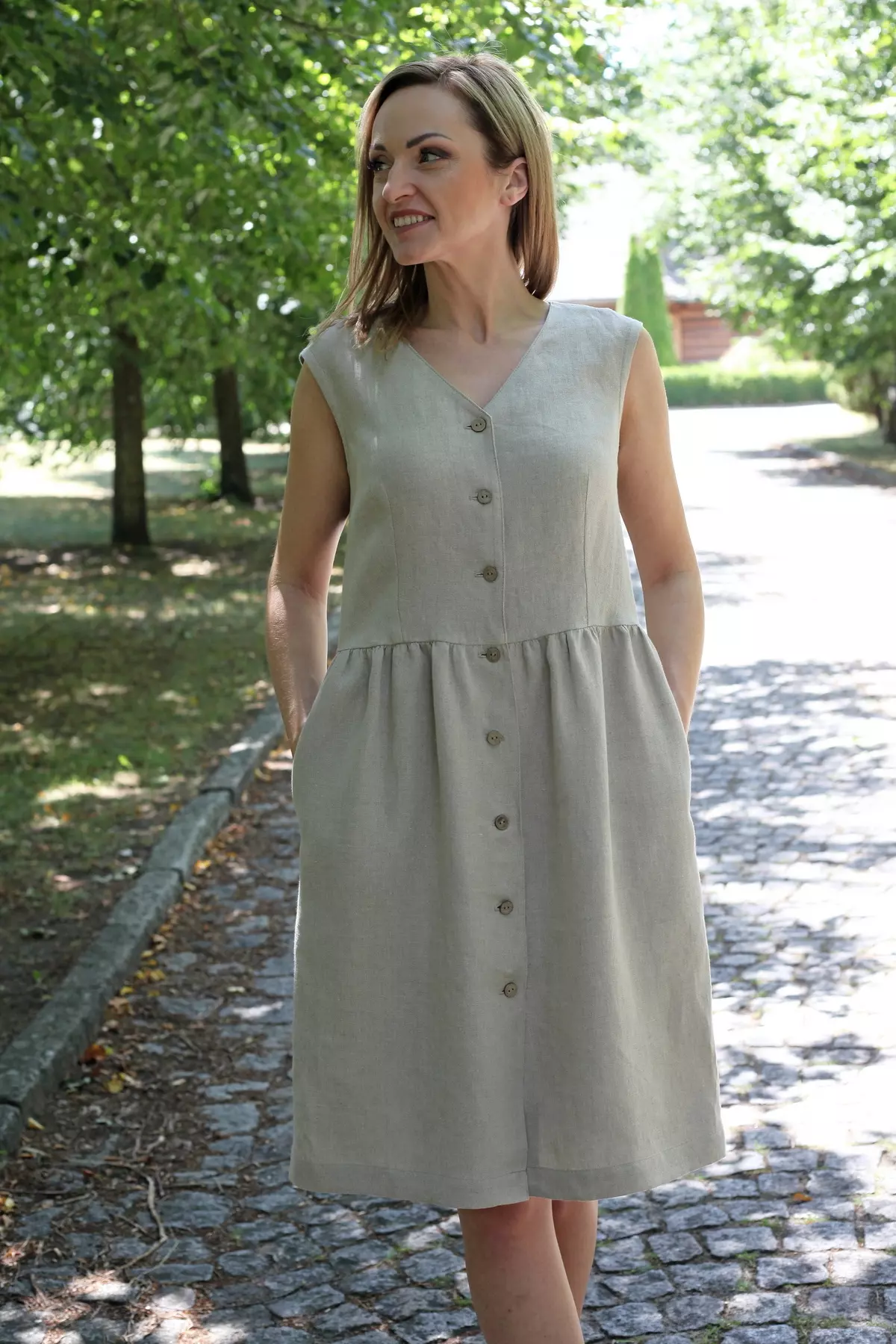 Modelka w beżowej sukience Lucy z lnu uśmiecha się, stojąc na alejce wśród drzew.