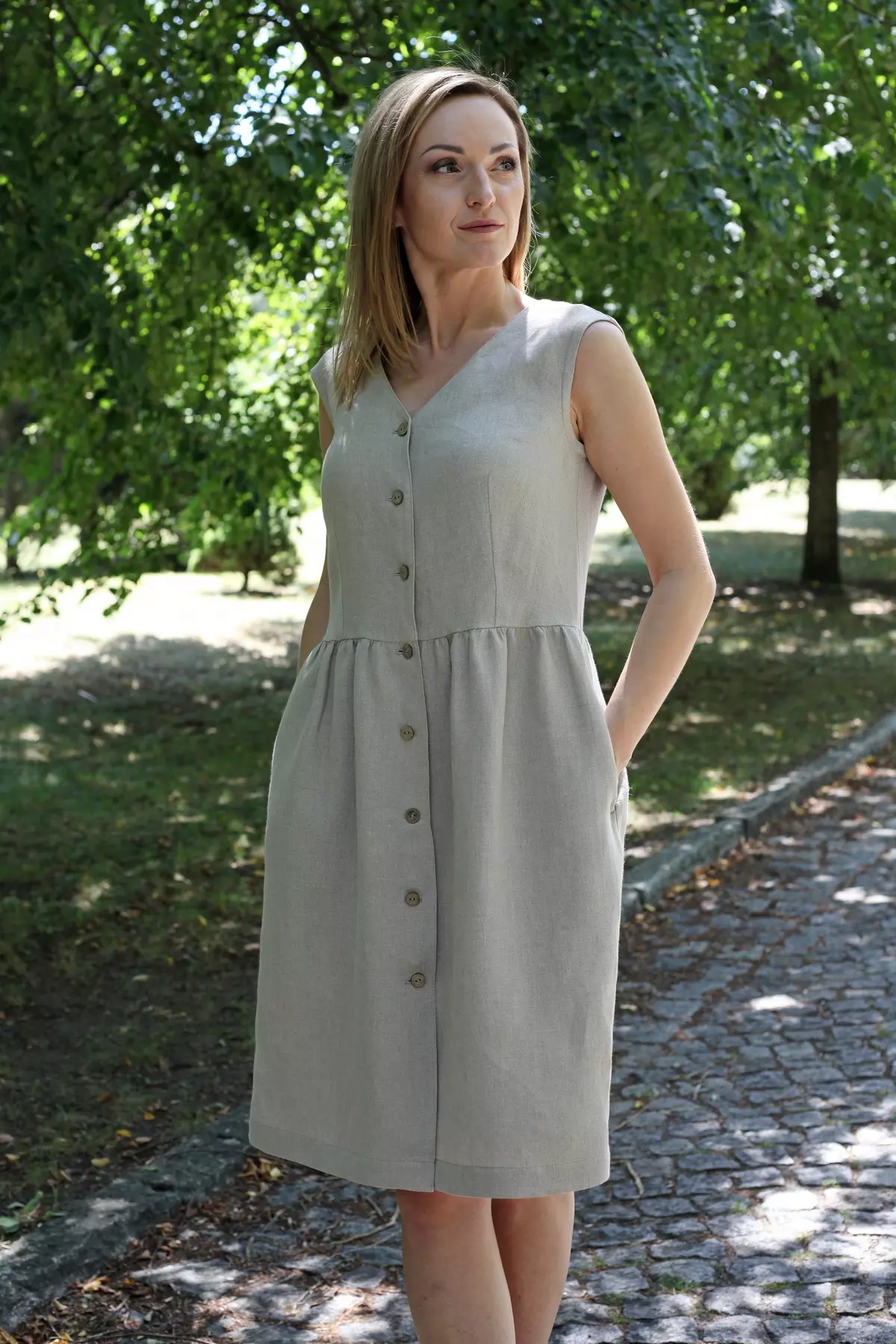 Modelka w lnianej sukience Lucy stoi w parku, z rękami w kieszeniach, wśród drzew i światła słonecznego.