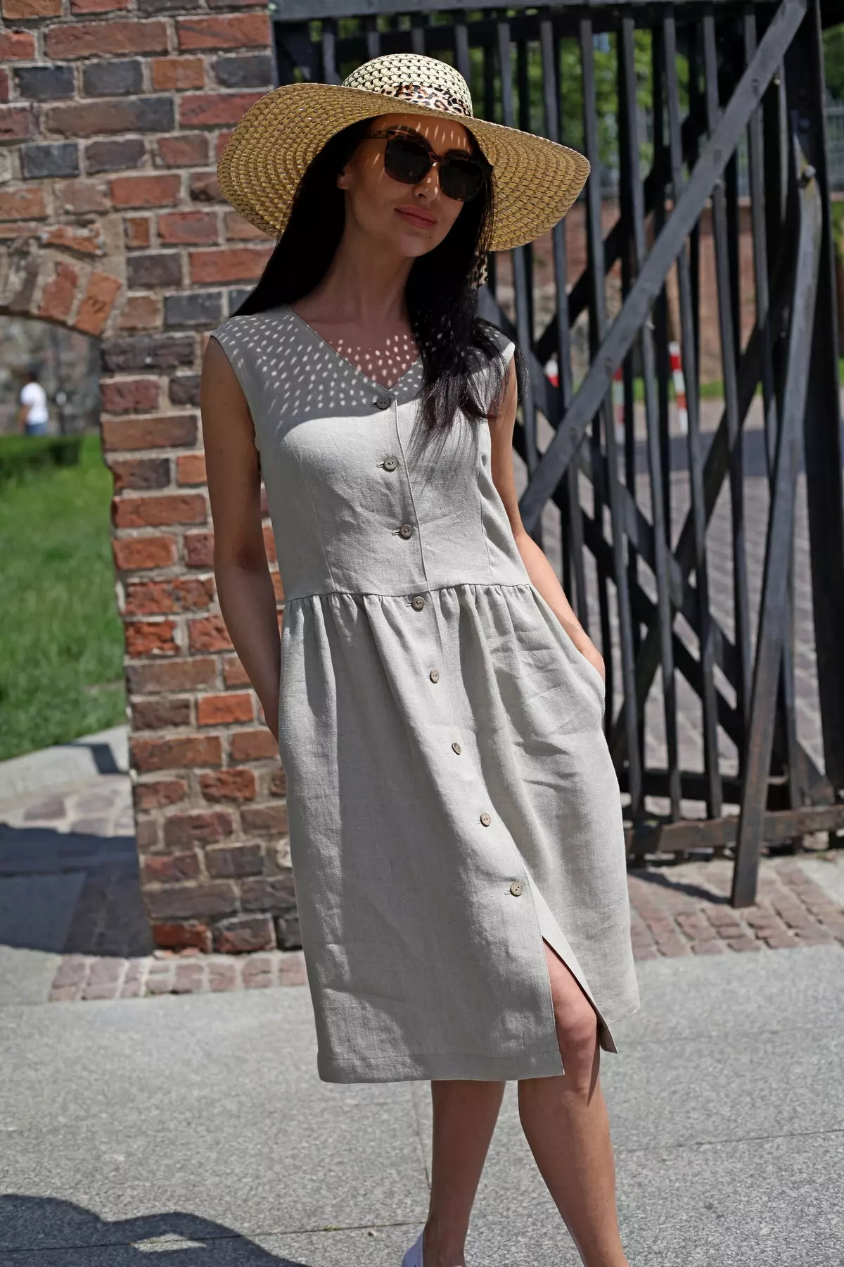 Modelka w jasnobeżowej lnianej sukience Lucy stoi przy ceglanej bramie, w słomkowym kapeluszu i okularach przeciwsłonecznych.