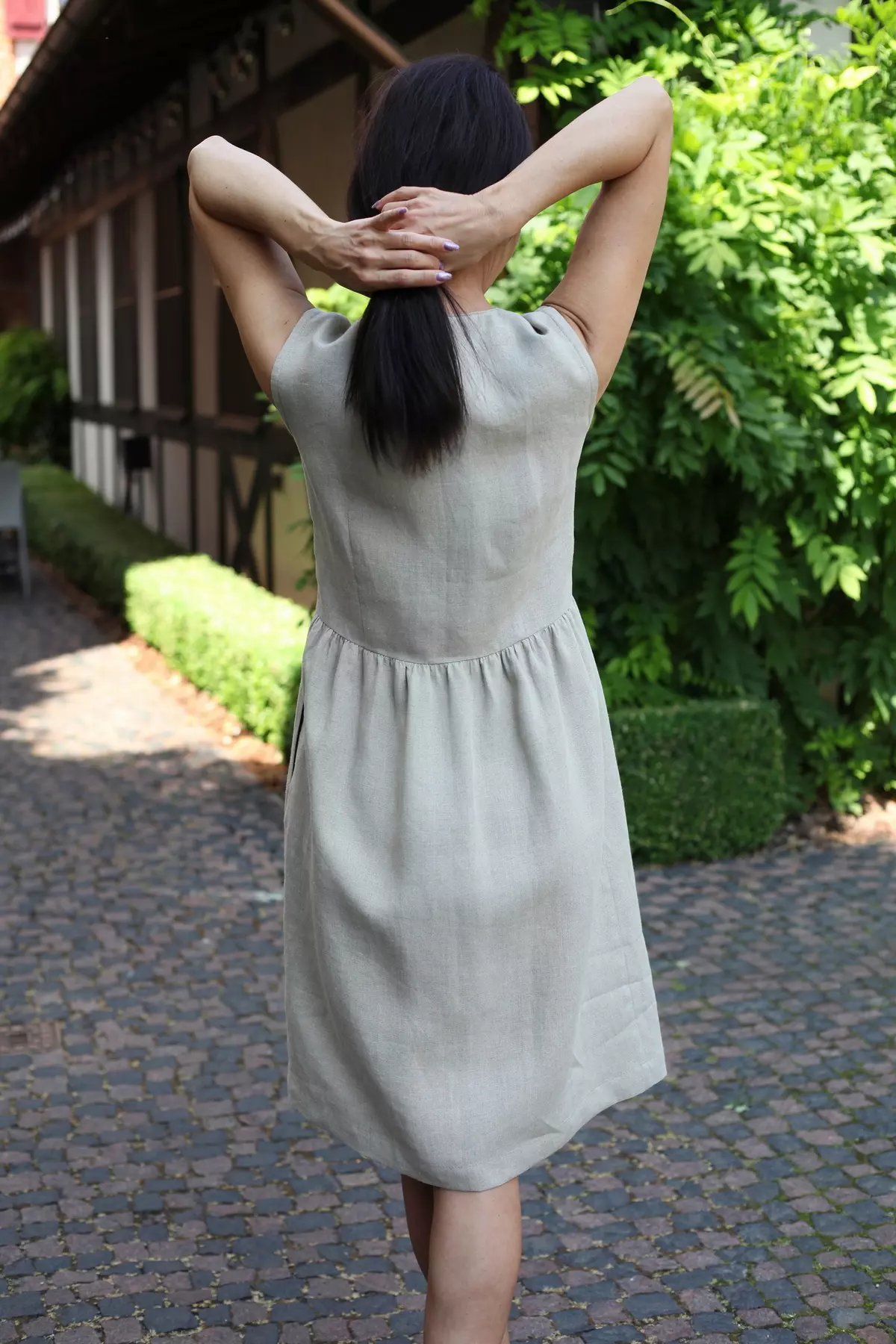 Modelka w lnianej sukience Lucy widziana od tyłu, z rękami splecionymi za głową, stoi w ogrodzie przy brukowanej ścieżce.