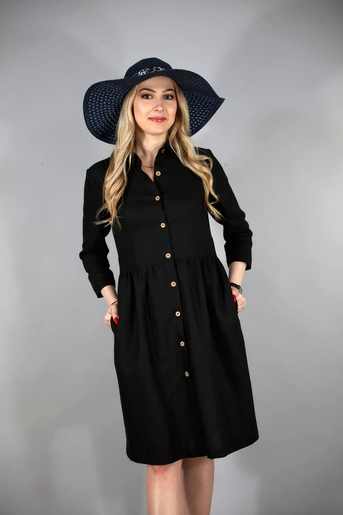Portret modelki w kapeluszu i czarnej sukience Lena. Modelka uśmiecha się, trzymając jedną rękę na biodrze. Sukienka jest lekko dopasowana w górnej części i rozszerza się ku dołowi, nadając sylwetce kształt klepsydry.