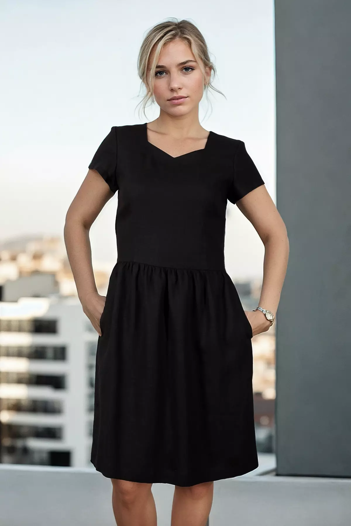 Kobieta stoi przodem do kamery z obiema rękami włożonymi do kieszeni sukienki, co lekko rozszerza spódnicę na boki, pokazując jej objętość. Sukienka jest czarna, matowa, sięgająca przed kolana. Modelka ma lekki uśmiech, a tło za nią to rozmyte budynki miejskie w dziennym świetle.