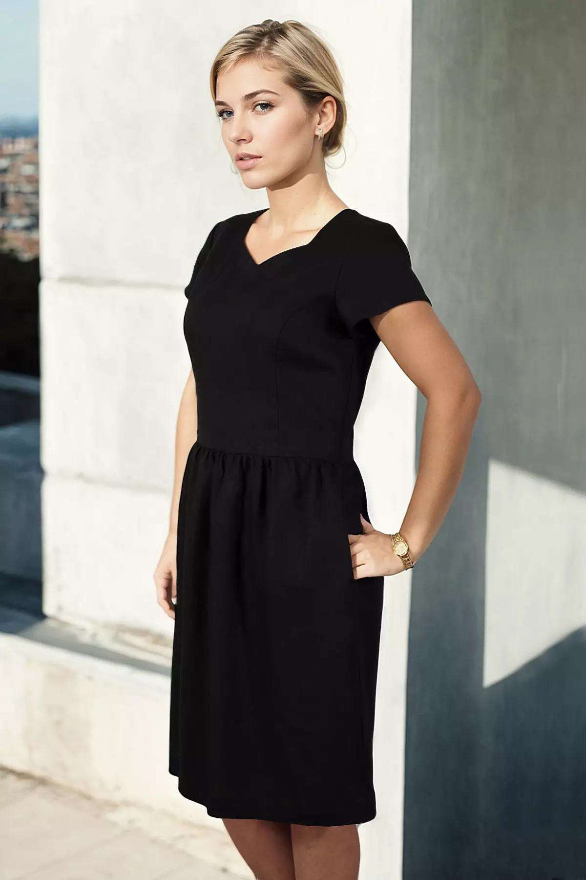 Ujęcie w półprofilu, od kolan w górę. Modelka patrzy w dal, w lewą stronę. Zdjęcie dobrze ukazuje dopasowanie góry sukienki do sylwetki oraz kształt krótkiego rękawka. Lewa ręka modelki spoczywa w kieszeni, na nadgarstku widoczny jest złoty zegarek. Światło słoneczne pada z boku, podkreślając rysy twarzy i fakturę materiału.