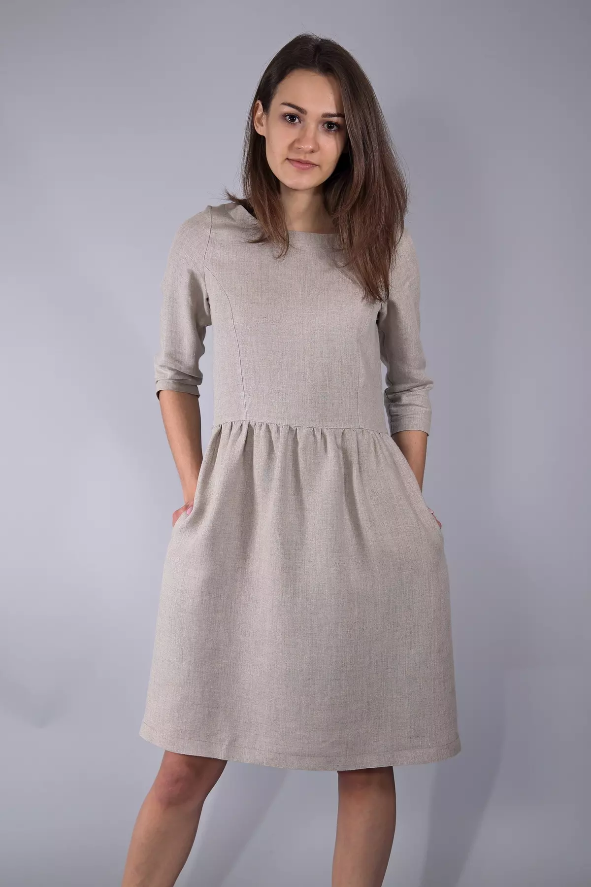 Brunetka stoi w studiu na jasnym tle. Ma na sobie sukienkę Zoja w kolorze naturalnego lnu. Dłonie trzyma w kieszeniach bocznych, co lekko rozszerza spódnicę. Sukienka jest minimalistyczna i elegancka.