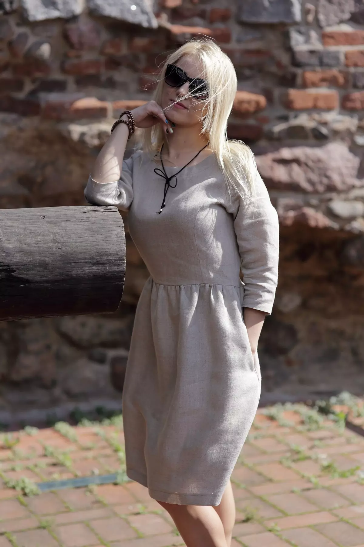 Modelka opiera się łokciem o grubą, drewnianą belkę. Wiatr rozwiewa jej blond włosy. Sukienka Zoja w kolorze naturalnym wygląda tu bardzo swobodnie i komfortowo. Widać długość rękawa 3/4.