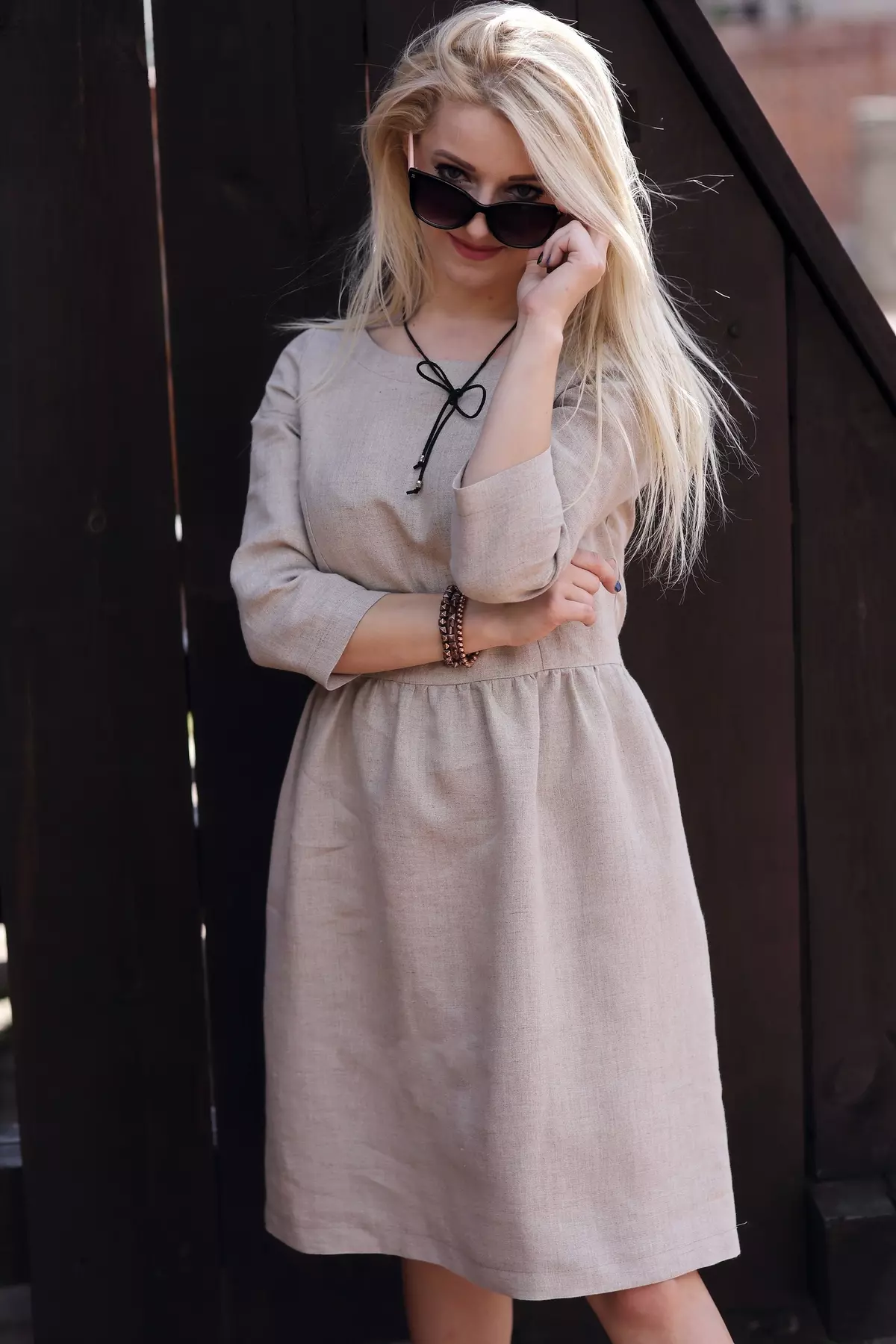 Zbliżenie na modelkę. Blondynka w ciemnych okularach uśmiecha się, dotykając oprawki. Widać fakturę niebarwionego lnu sukienki Zoja. Dekolt jest prosty, bez kołnierzyka, ozdobiony rzemykiem z wisiorkiem.