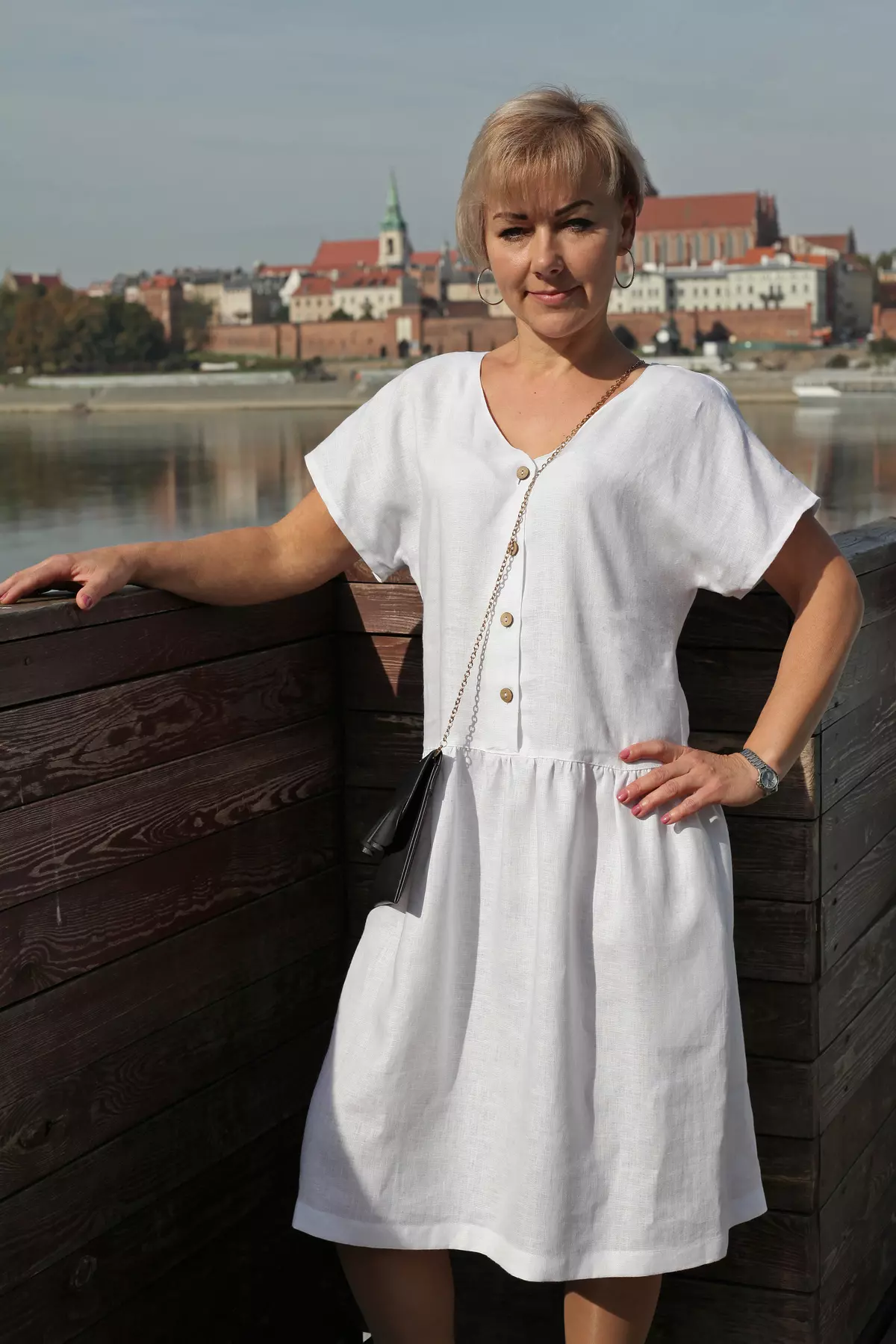 Kobieta o krótkich włosach stoi na drewnianym pomoście na tle panoramy miasta i rzeki. Ma na sobie białą sukienkę Rita i małą torebkę przewieszoną przez ramię. Słońce oświetla biały materiał, podkreślając jego fakturę.