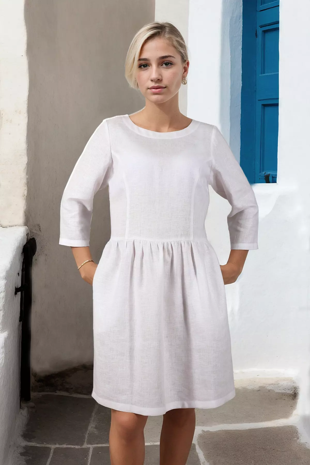 Modelka stoi na tle białej ściany z niebieską okiennicą. Trzyma obie dłonie w kieszeniach ukrytych w bocznych szwach sukienki, co lekko rozszerza spódnicę na biodrach. Biały len wygląda na przewiewny i lekki, kontrastując z letnią opalenizną modelki.