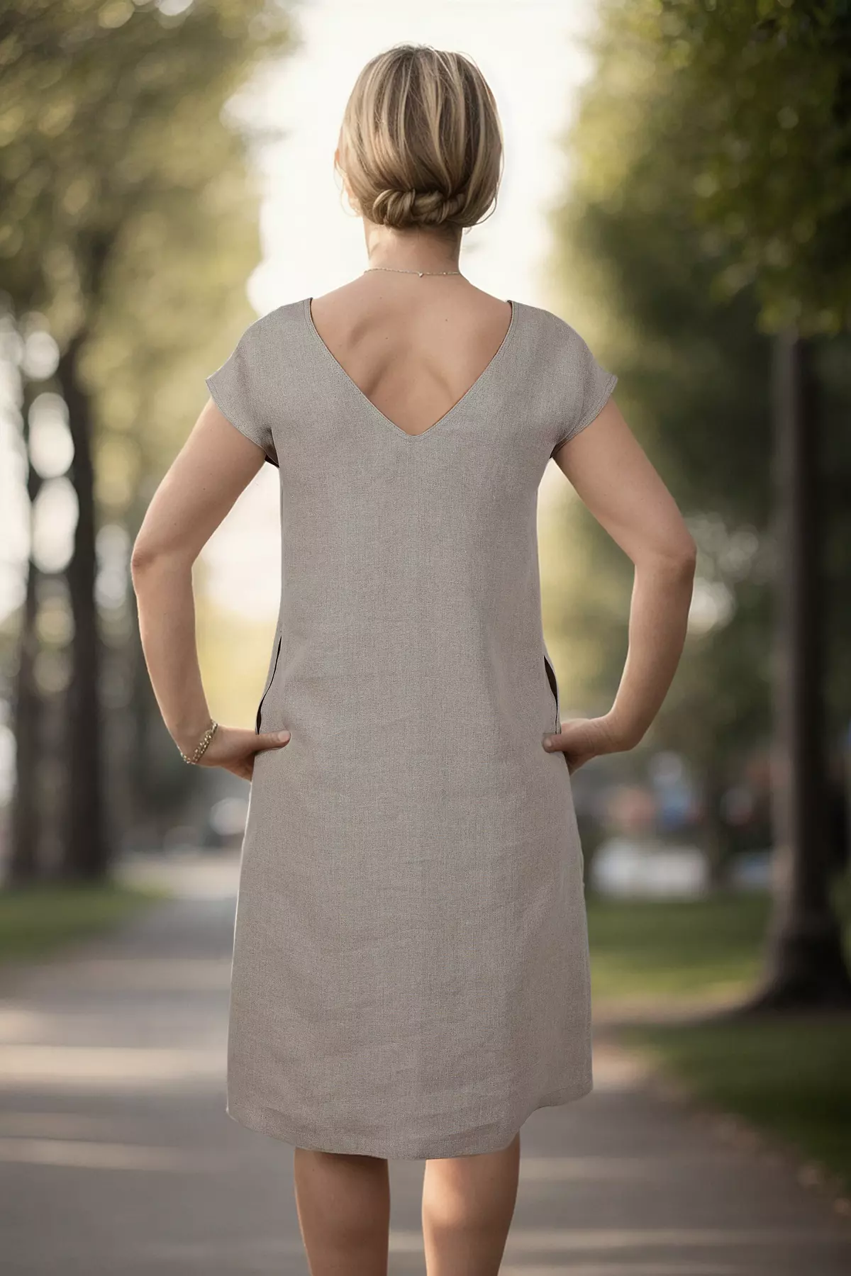 Modelka stoi tyłem w parkowej alejce. Zdjęcie ukazuje tył sukienki w kolorze naturalnego lnu. Kluczowym elementem jest głęboki dekolt w kształcie litery V, który odsłania górną część pleców. Sukienka ma luźny krój.