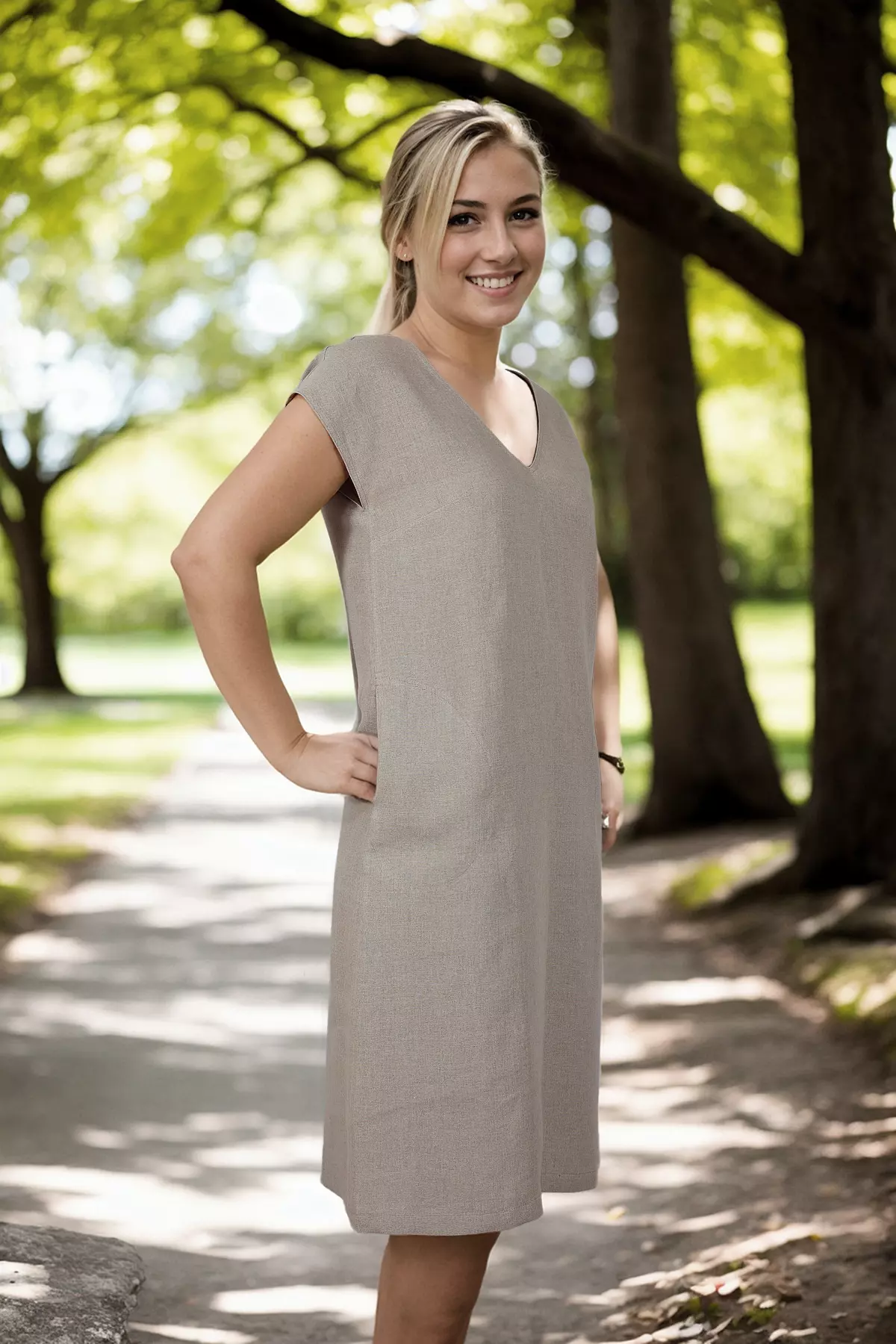 Uśmiechnięta modelka w półprofilu. Stoi w cieniu drzew. Ma na sobie beżową sukienkę lnianą. Widać, jak materiał swobodnie układa się na sylwetce. Ręka modelki jest oparta o biodro, przy kieszeni.