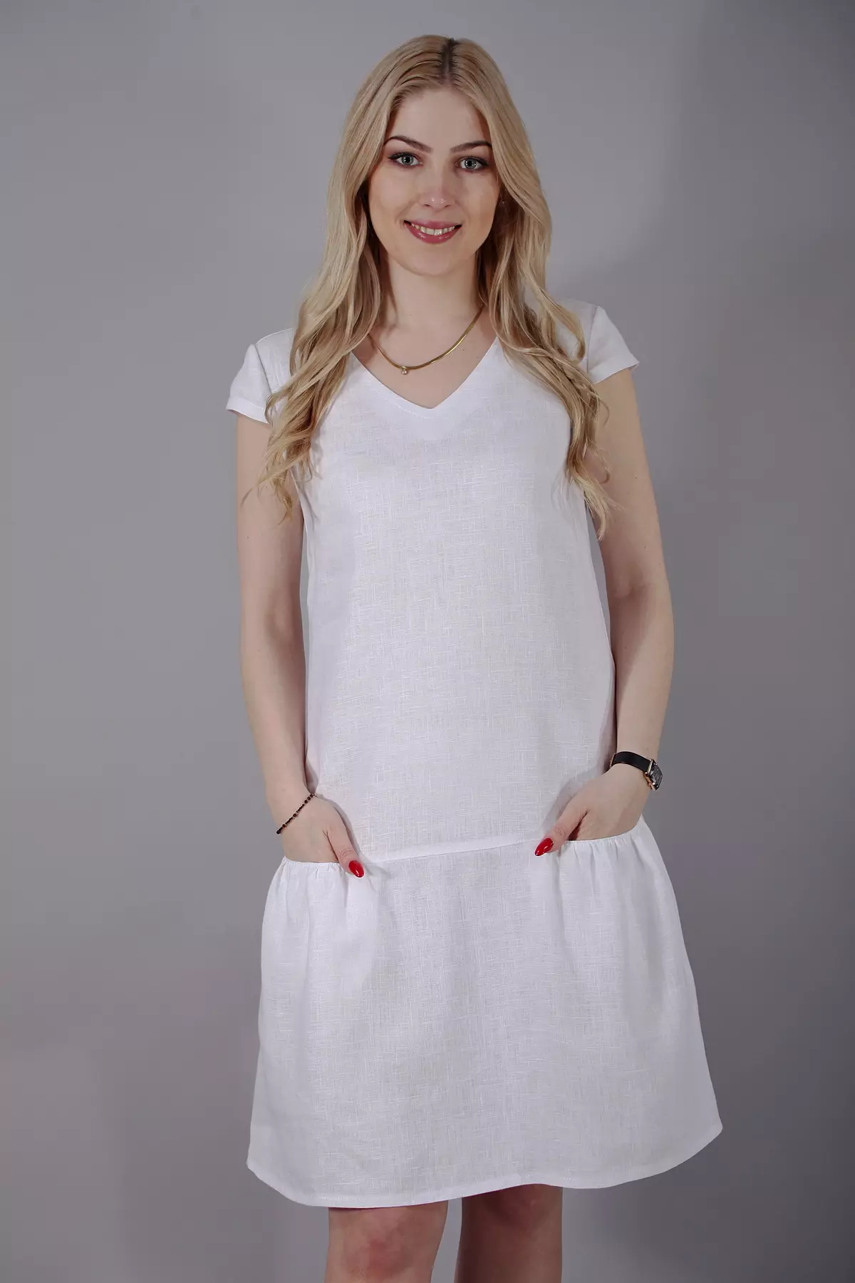Portret uśmiechniętej blondynki w białej sukience. Ujęcie ukazuje dekolt w kształcie litery V oraz luźno opadający materiał, który nie opina sylwetki. Ręce modelki są schowane w kieszeniach, podkreślając swobodę fasonu.