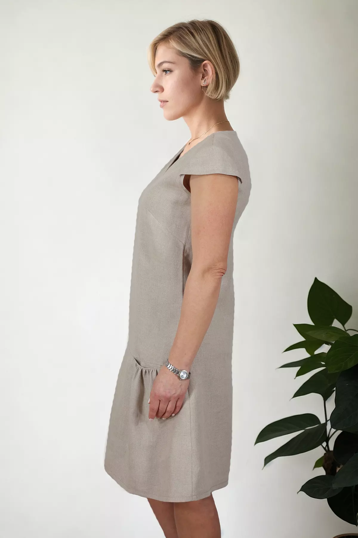 Widok z boku na lnianą sukienkę Dora. Jasnobeżowa sukienka o kroju trapezowym (litera A) sięgająca przed kolano. Wyraźnie widoczna głębokość marszczonej kieszeni bocznej oraz naturalna sztywność lnianej tkaniny.