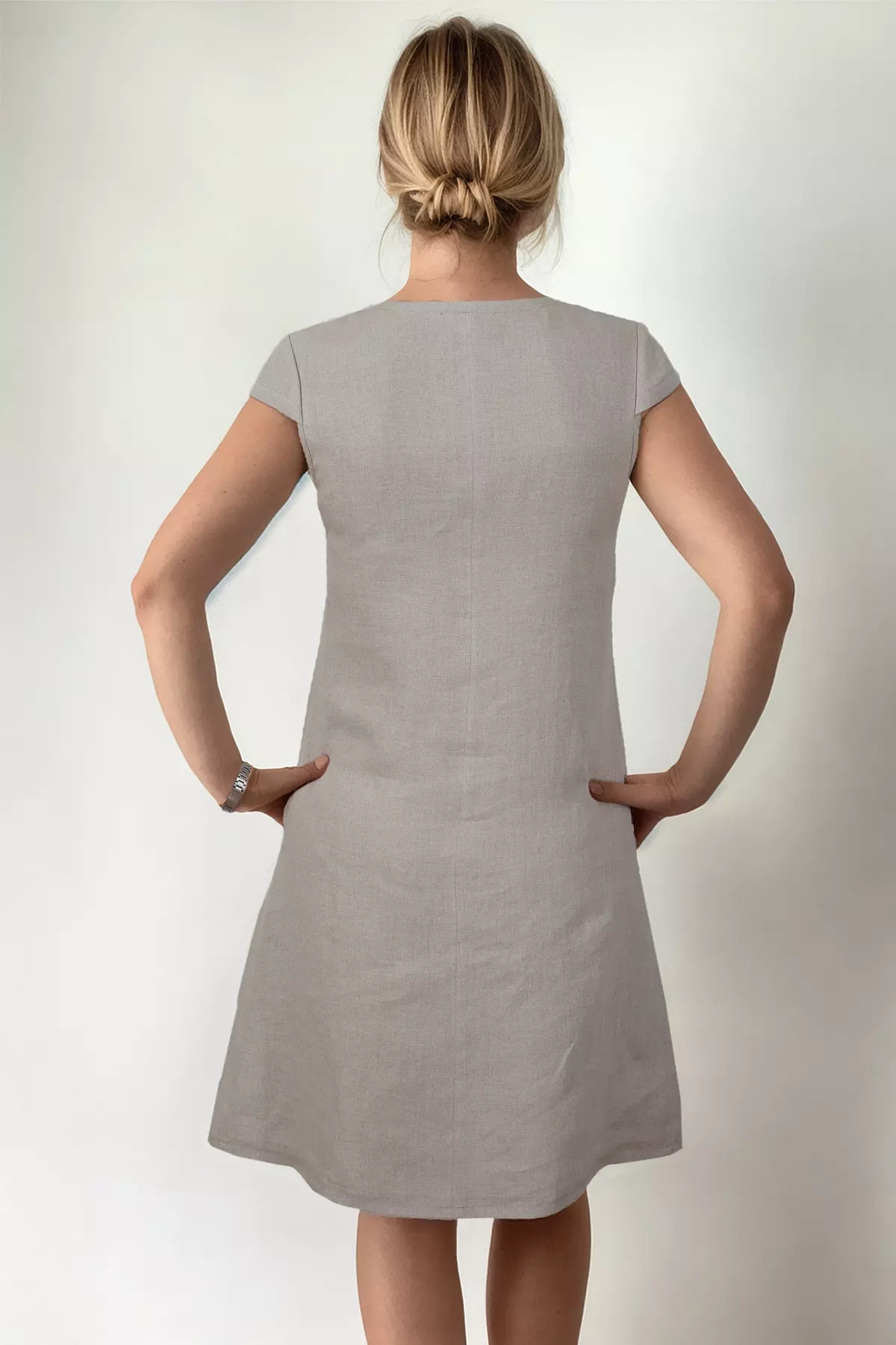 Tył lnianej sukienki Dora w kolorze naturalnym. Zdjęcie pokazuje prosty krój pleców z pionowym przeszyciem przez środek oraz krótkie, dopasowane rękawki. Modelka ma włosy upięte w niski kok.