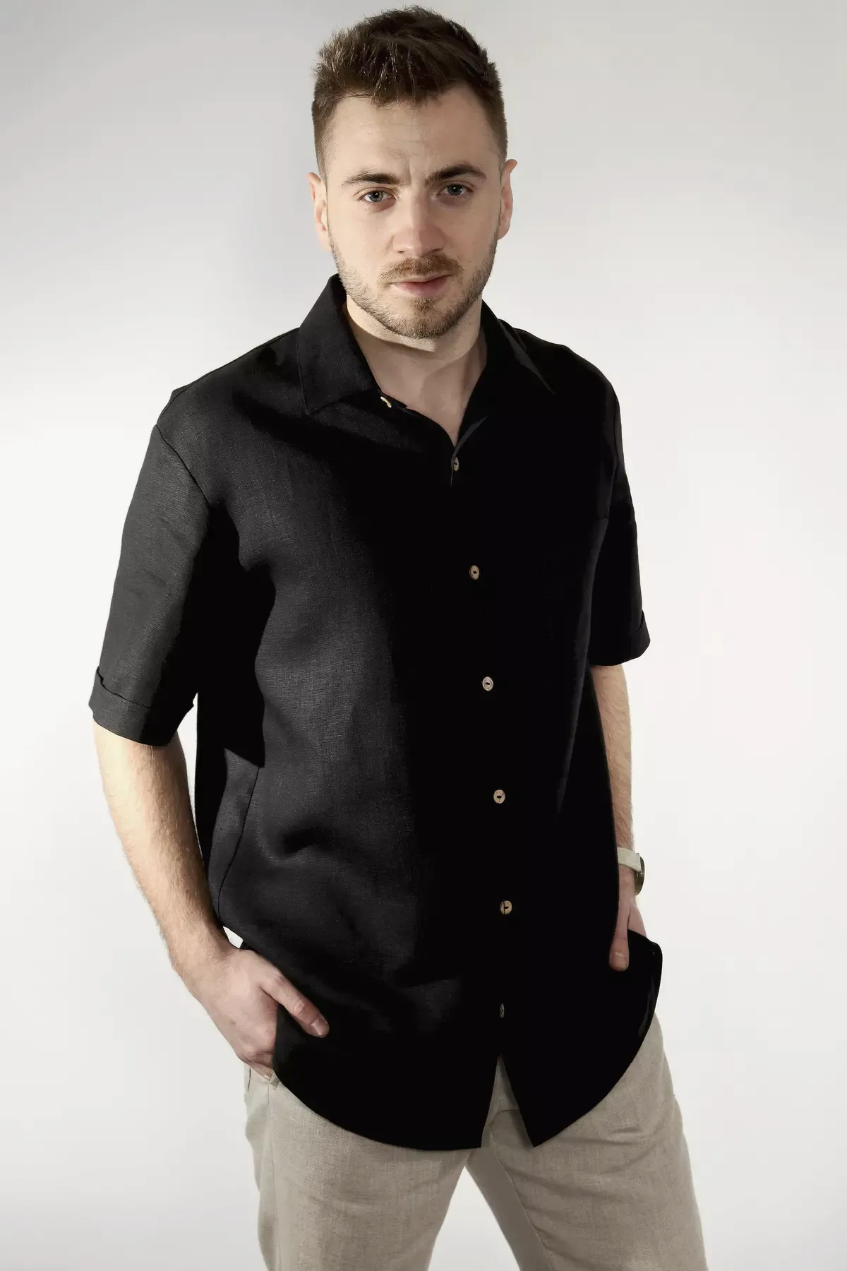 Mężczyzna stoi w półprofilu. Ma na sobie czarną koszulę lnianą Goran z krótkim rękawem i kołnierzykiem. Ręce trzyma w kieszeniach beżowych spodni. Koszula zapinana jest na jasne, kontrastowe guziki.