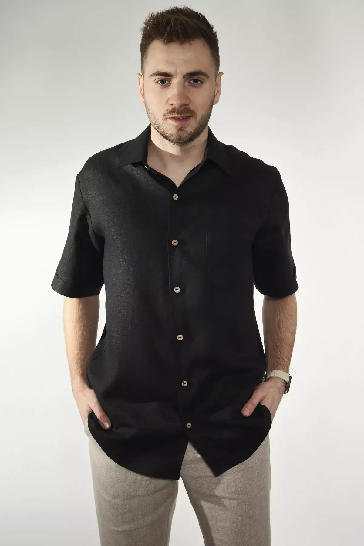 Mężczyzna stoi prosto na białym tle. Prezentuje czarną koszulę Goran. Widać klasyczny kołnierzyk, kieszonkę na piersi i jasne guziki. Koszula jest luźno wypuszczona na jasne spodnie. Ręce w kieszeniach.