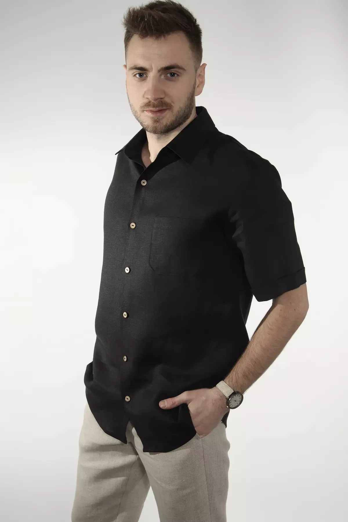 Mężczyzna stoi w półprofilu. Ma na sobie czarną koszulę lnianą Goran z krótkim rękawem i kołnierzykiem. Ręce trzyma w kieszeniach beżowych spodni. Koszula zapinana jest na jasne, kontrastowe guziki.