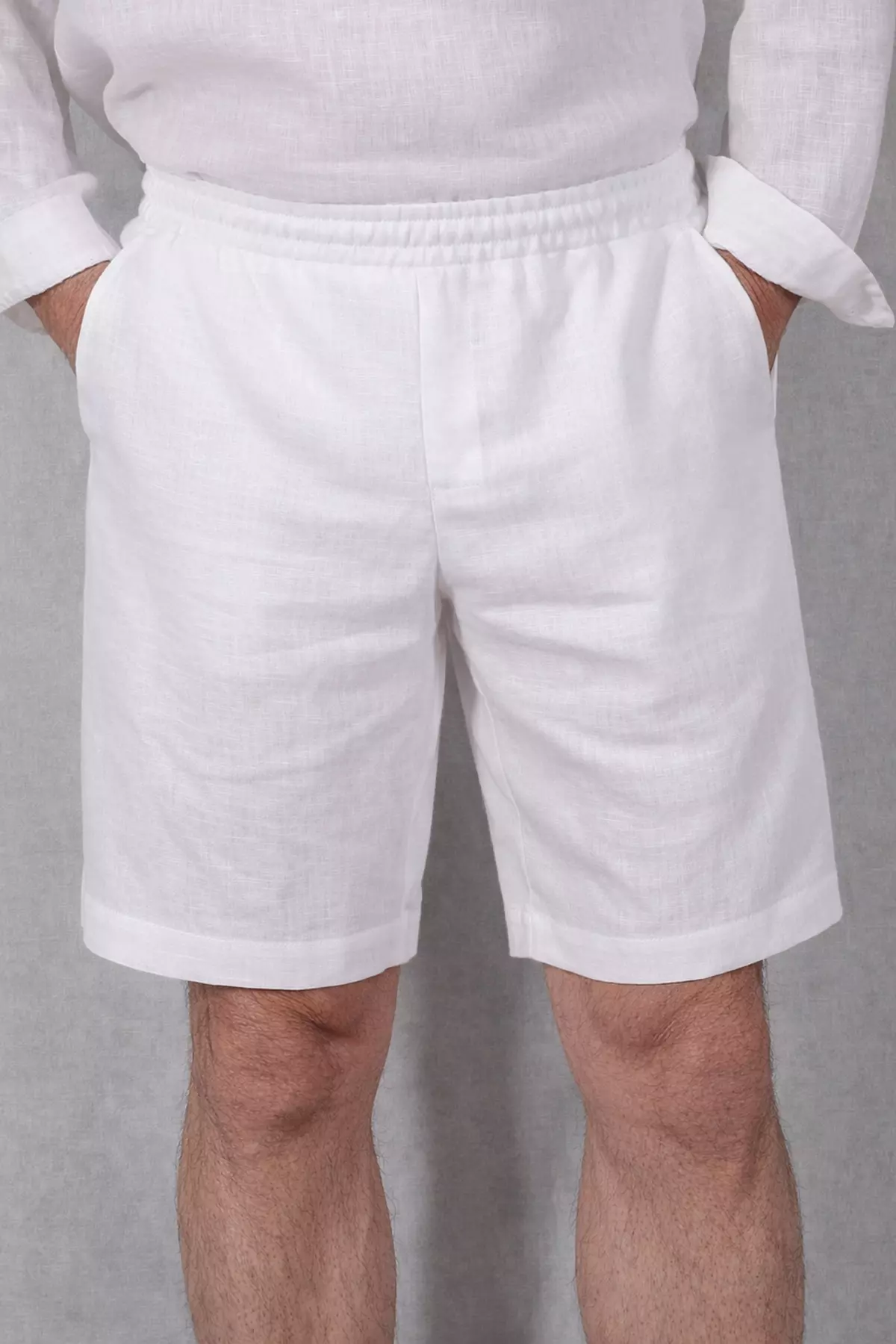 Zdjęcie od pasa w dół przedstawiające mężczyznę w śnieżnobiałych, lnianych szortach. Model Alex ma prosty, luźny krój sięgający przed kolano. Mężczyzna trzyma dłonie w bocznych kieszeniach. W pasie widoczna jest marszczona guma.