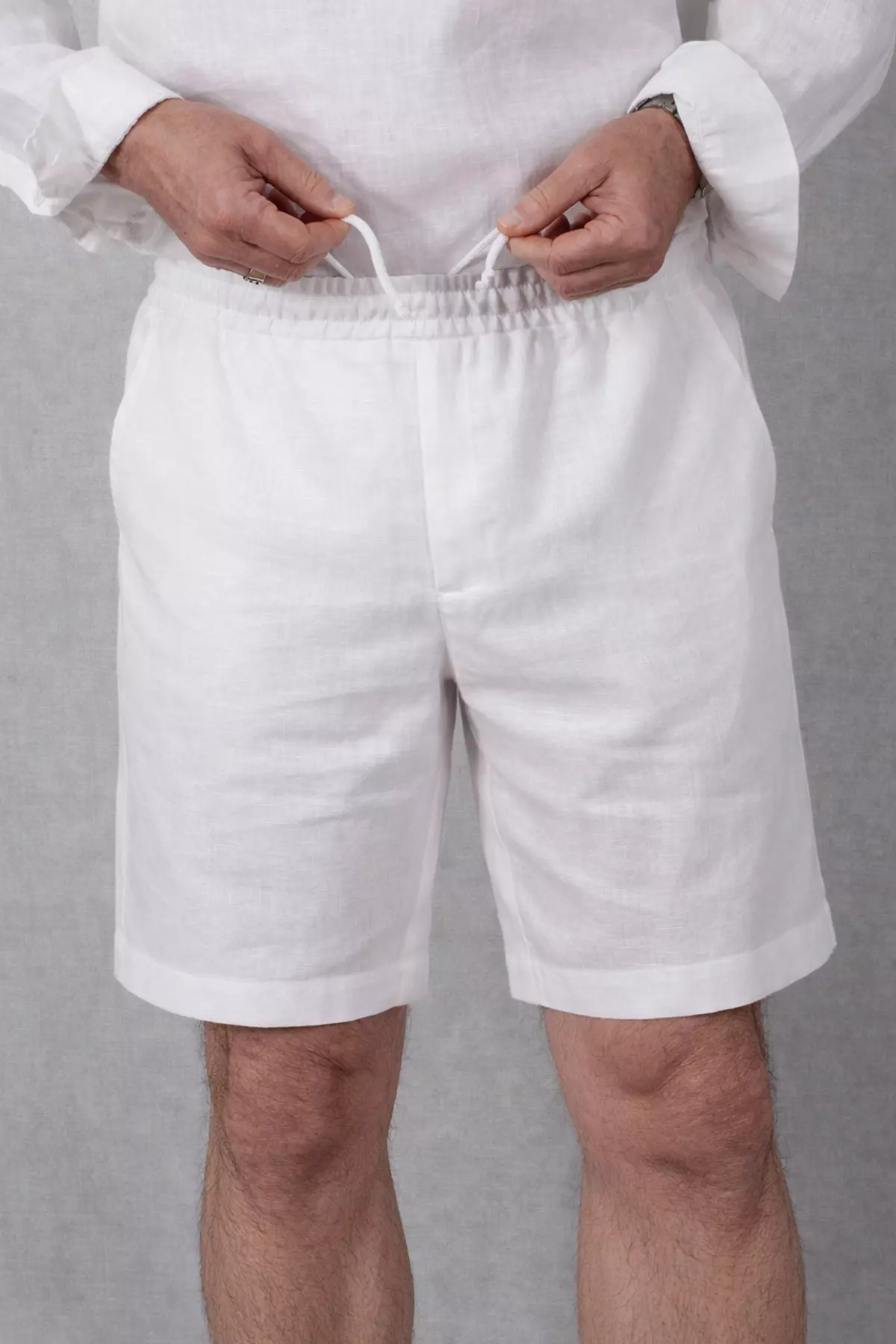 Zbliżenie na górną część białych szortów. Dłonie mężczyzny wiążą biały, bawełniany sznurek w pasie. Wyraźnie widać fakturę lnu oraz elastyczne marszczenie gumy. Na środku widoczne przeszycie imitujące rozporek.