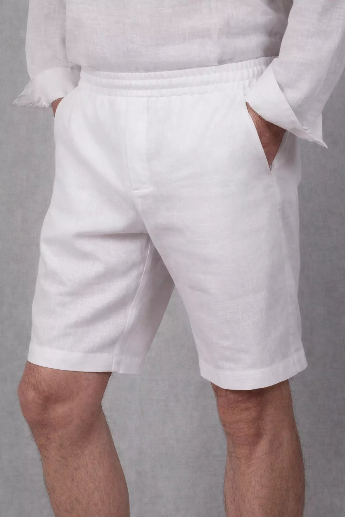 Widok z boku na białe spodenki lniane. Mężczyzna stoi w lekkim rozkroku, lewą dłoń trzyma w kieszeni, co lekko napina materiał. Widać, że tkanina jest przewiewna i lekka. Góra stylizacji to biała lniana koszula.