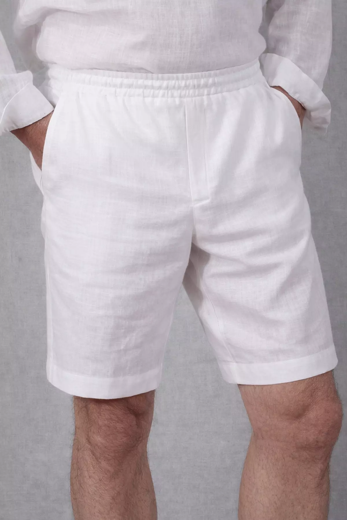 Ujęcie pod kątem (półprofil). Mężczyzna trzyma dłonie w kieszeniach. Zdjęcie ukazuje strukturę białego lnu oraz swobodę, jaką daje krój Regular Fit. Widać wyraźnie linię rozporka.