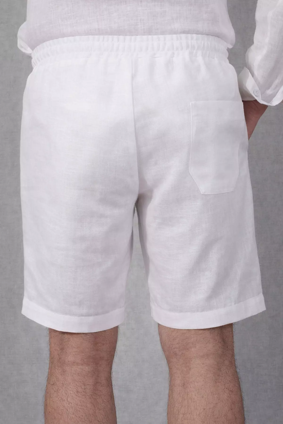 Widok szortów Alex od tyłu. Na prawym pośladku naszyta jest jedna, otwarta kieszeń o kwadratowym kształcie. Biały materiał kontrastuje z lekko opaloną skórą nóg modela. W pasie widoczne gęste marszczenie gumy.