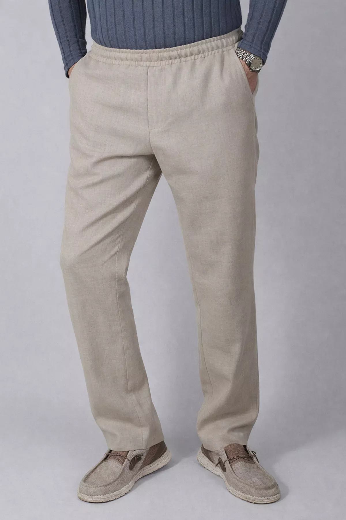 Mężczyzna w lnianych spodniach ujęty w lekkim półprofilu. Prawa noga jest wysunięta do przodu, jak w trakcie kroku. Zdjęcie ukazuje miękkość materiału i to, jak spodnie układają się na nogach. Kolor spodni to naturalny, jasny beż.