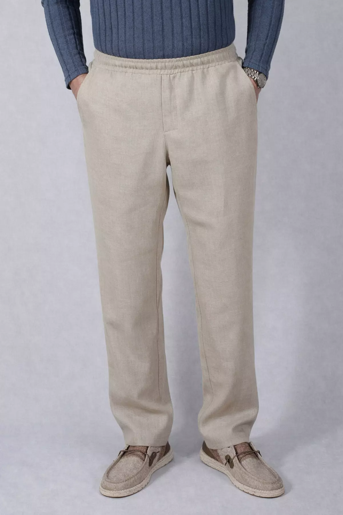 Zdjęcie całej sylwetki od pasa w dół. Mężczyzna stoi prosto, ubrany w długie, beżowe spodnie lniane o prostym kroju. Ręce trzyma w bocznych kieszeniach, co lekko napina materiał w biodrach. W pasie widoczna marszczona guma. Model ma na nogach beżowe mokasyny.