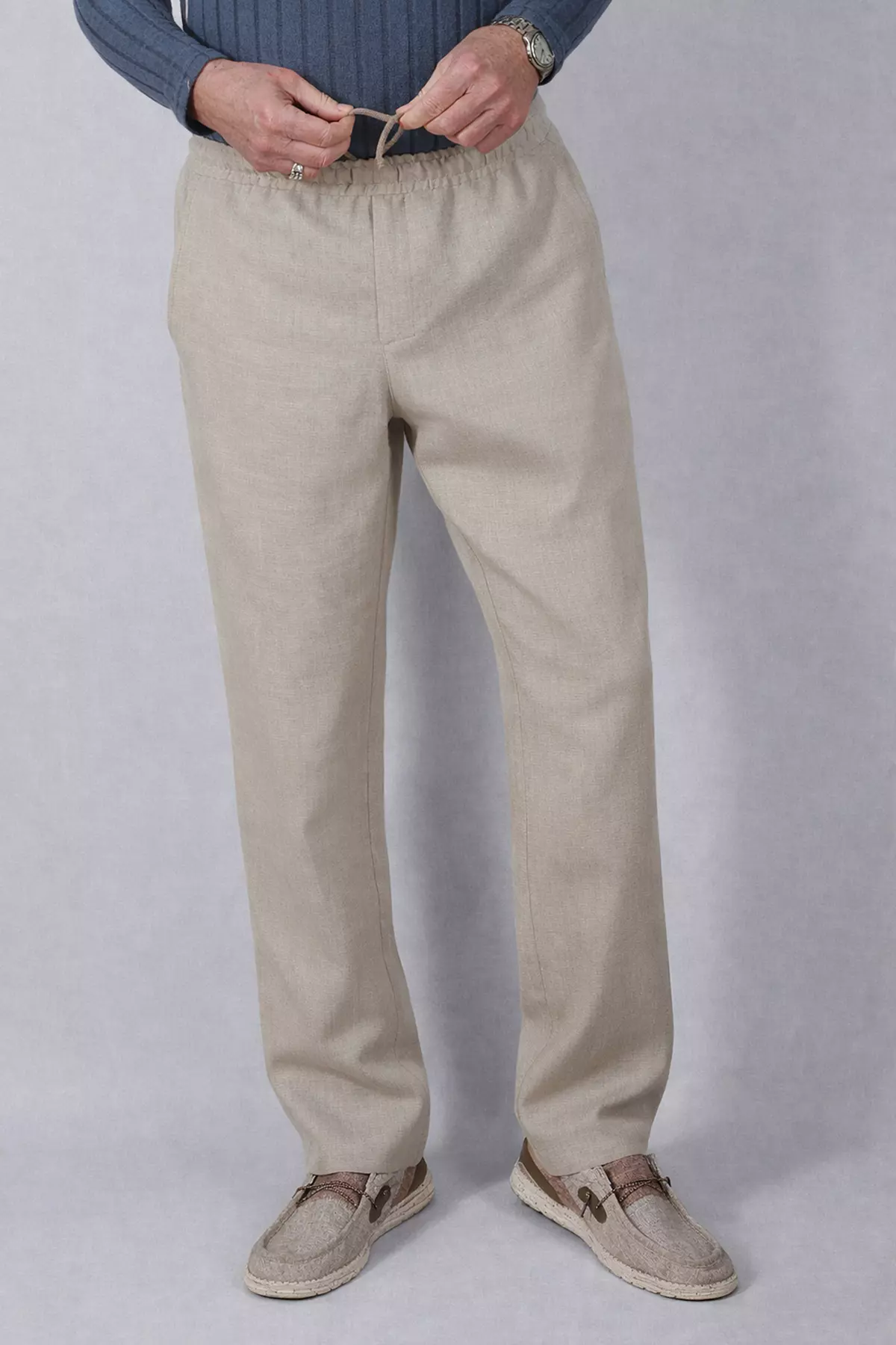 Zbliżenie na górną partię spodni (okolice pasa). Dłonie mężczyzny wiążą beżowy, bawełniany sznurek. Wyraźnie widać fakturę naturalnego lnu (splot włókien) oraz imitację rozporka na środku. Góra stroju to niebieski sweter o waflowej strukturze.