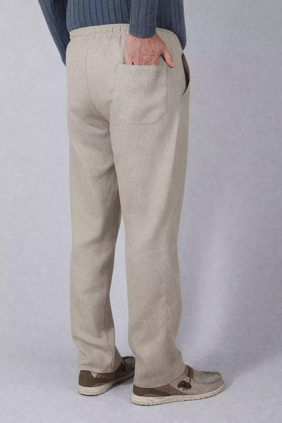 Widok spodni od tyłu. Mężczyzna stoi tyłem, prawa ręka wsunięta jest do tylnej, naszywanej kieszeni na prawym pośladku. Widać luźny krój nogawek i naturalne zagniecenia lnu w okolicach kolan.