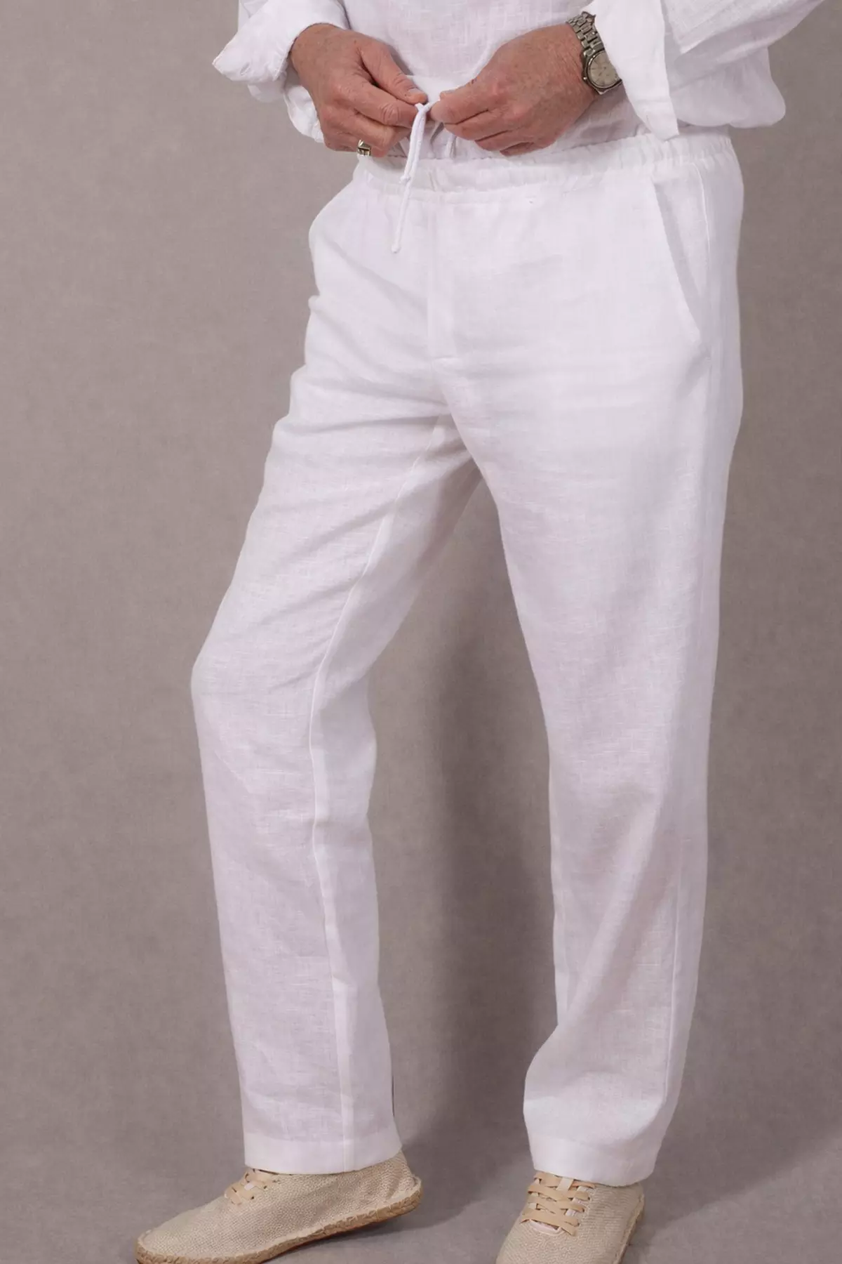Zbliżenie na górną część spodni w okolicach pasa. Mężczyzna trzyma w dłoniach końce białego sznurka, wiążąc go w pasie. Wyraźnie widać marszczenie elastycznej gumy oraz pionowe przeszycie imitujące linię rozporka. Materiał ma widoczną, naturalną strukturę lnu.