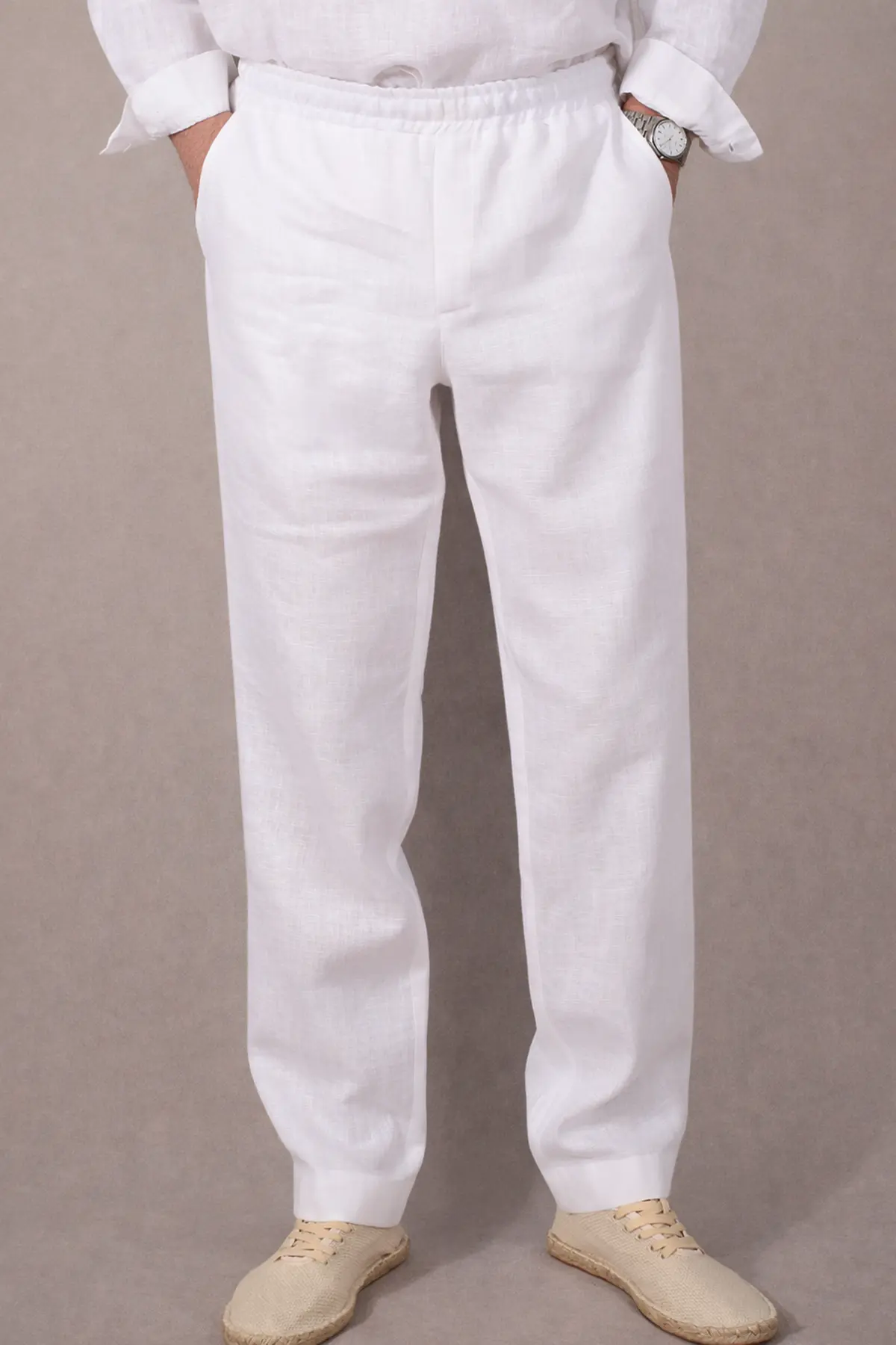 Mężczyzna stoi przodem, trzymając dłonie w bocznych kieszeniach spodni. Biały materiał lekko napina się na biodrach, ukazując wygodny krój. Spodnie zestawione są z białą lnianą koszulą, tworząc kompletny, letni zestaw (Total White Look).