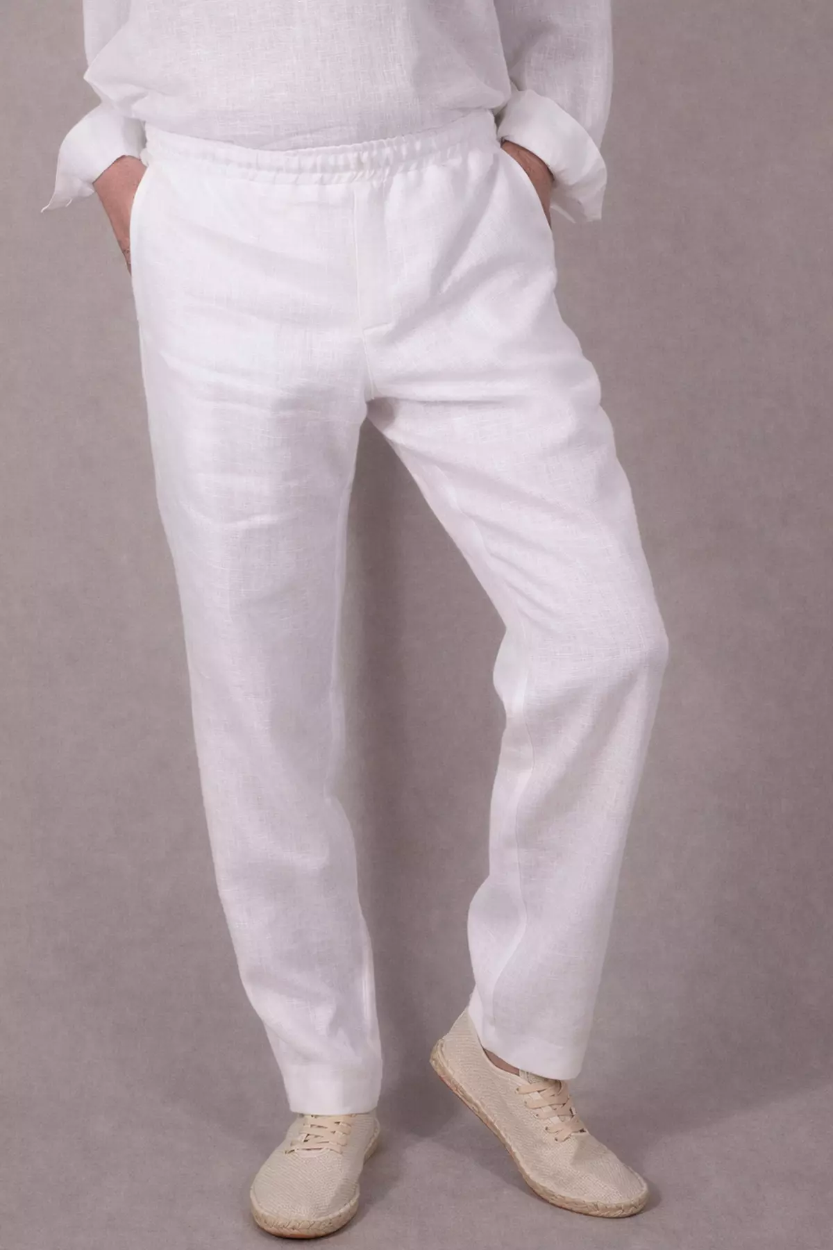 Ujęcie pod kątem. Mężczyzna stoi w swobodnej pozie. Widać detale konstrukcyjne spodni: boczne kieszenie, elastyczny pas oraz linię rozporka. Biały len delikatnie załamuje się na nogach, co świadczy o jego miękkości.